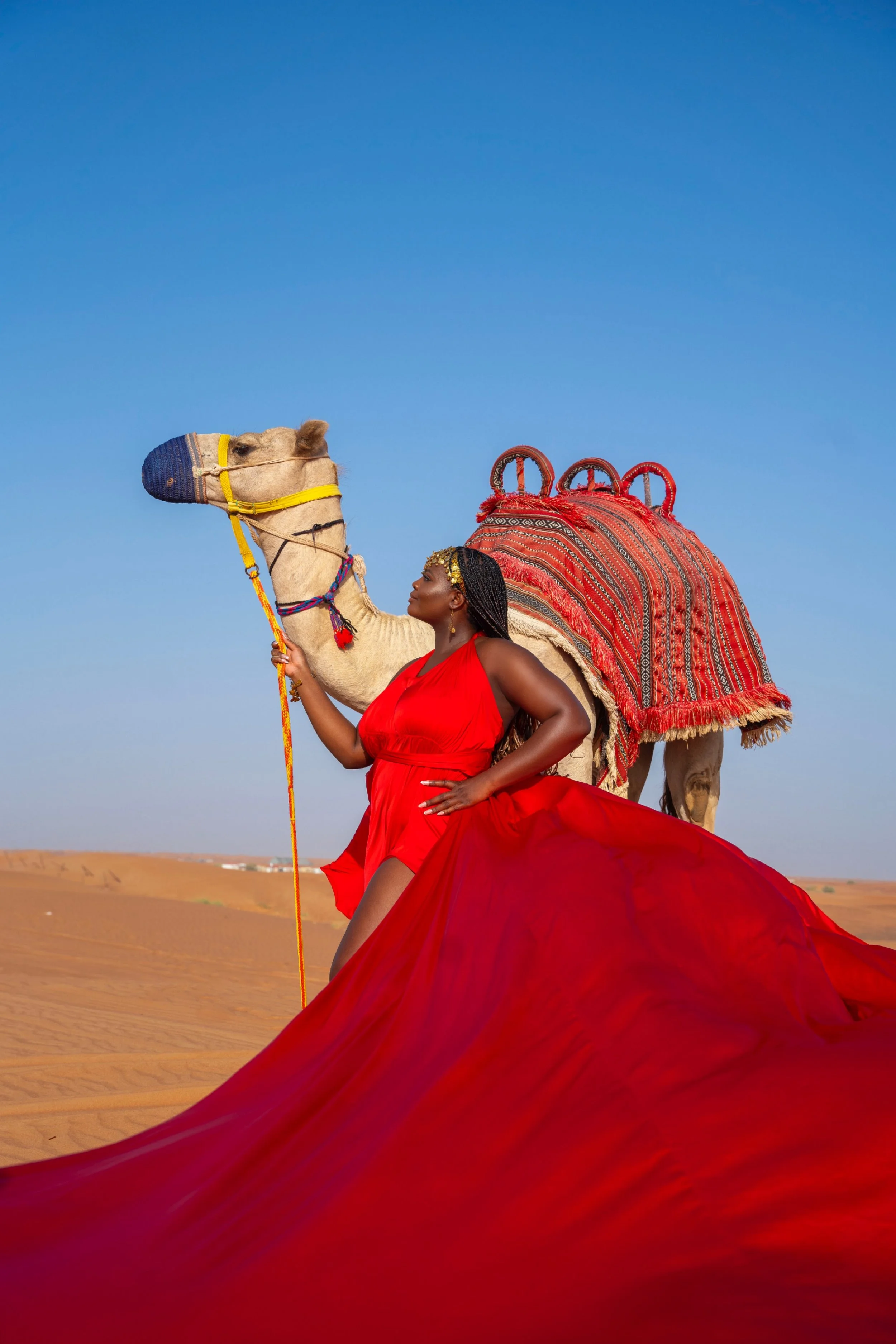 A woman in a red dress standing in a desert, holding a camel's reins, with the camel's decorated back visible behind her.