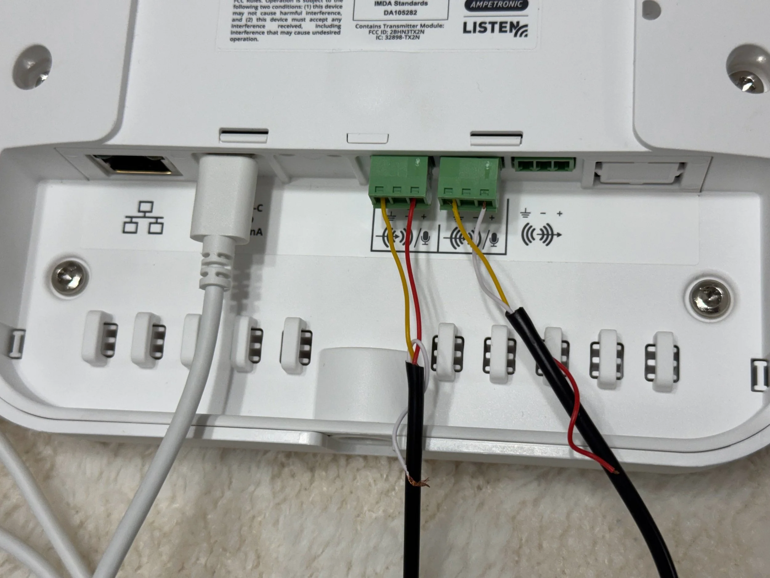 Close-up view of the bottom side of a white electronic device, showing various ports, wires, and labels, with a beige textured surface underneath.