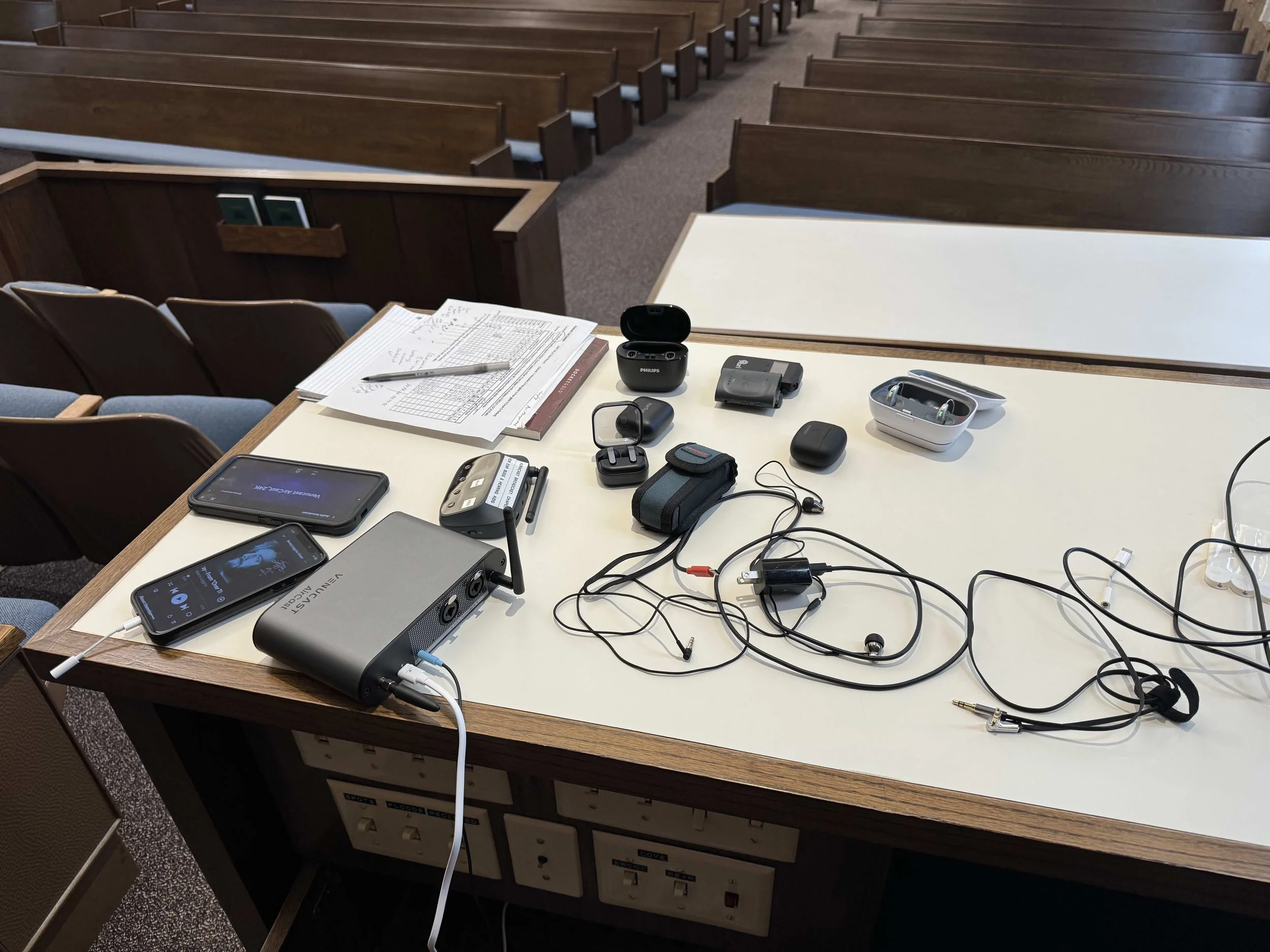 View of a church or lecture hall with wooden pews and a white table at the front, which has various electronic devices, papers, and objects including earbuds, a pen, and chargers.