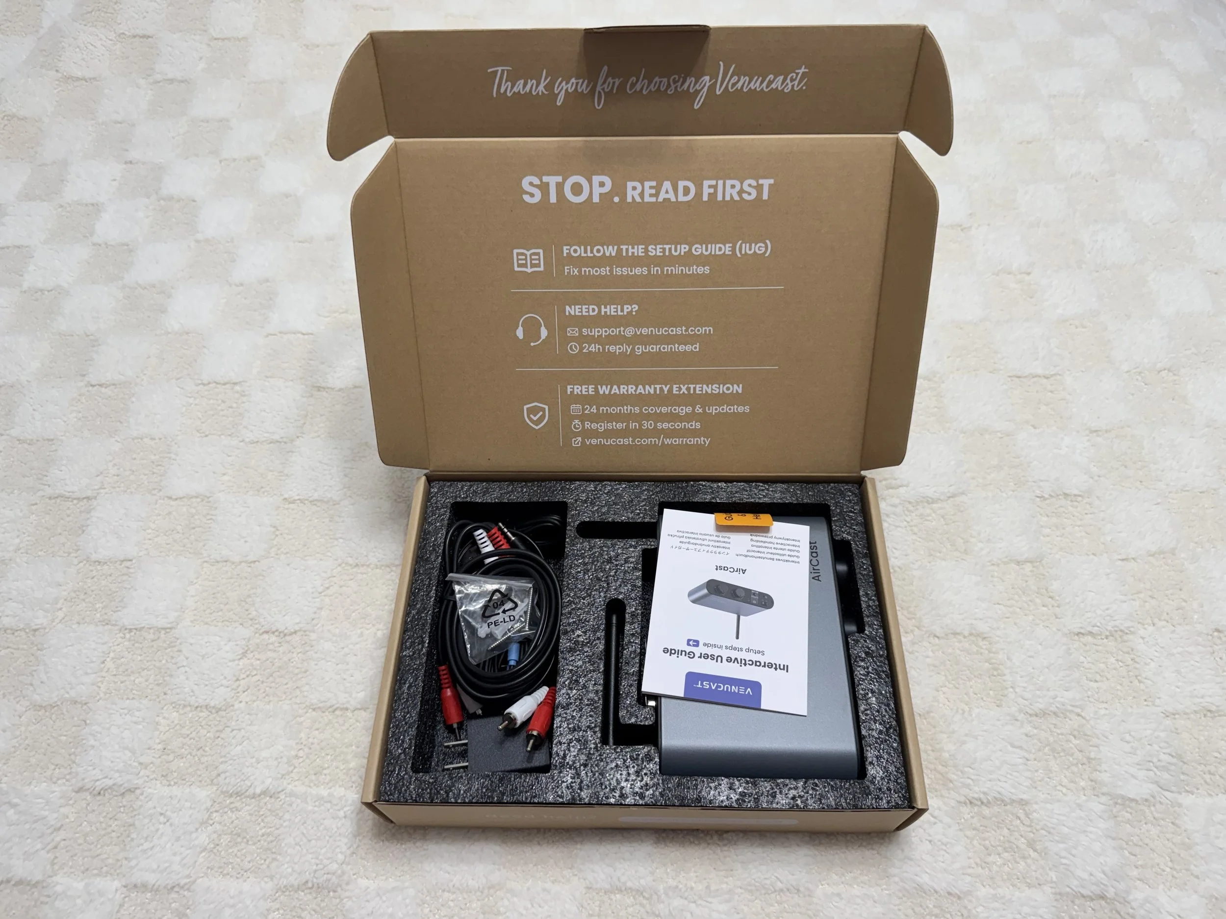 Open cardboard box containing electronic accessories, including cables and user guide, on a textured surface. The box lid has instructions for setup and support contact information.