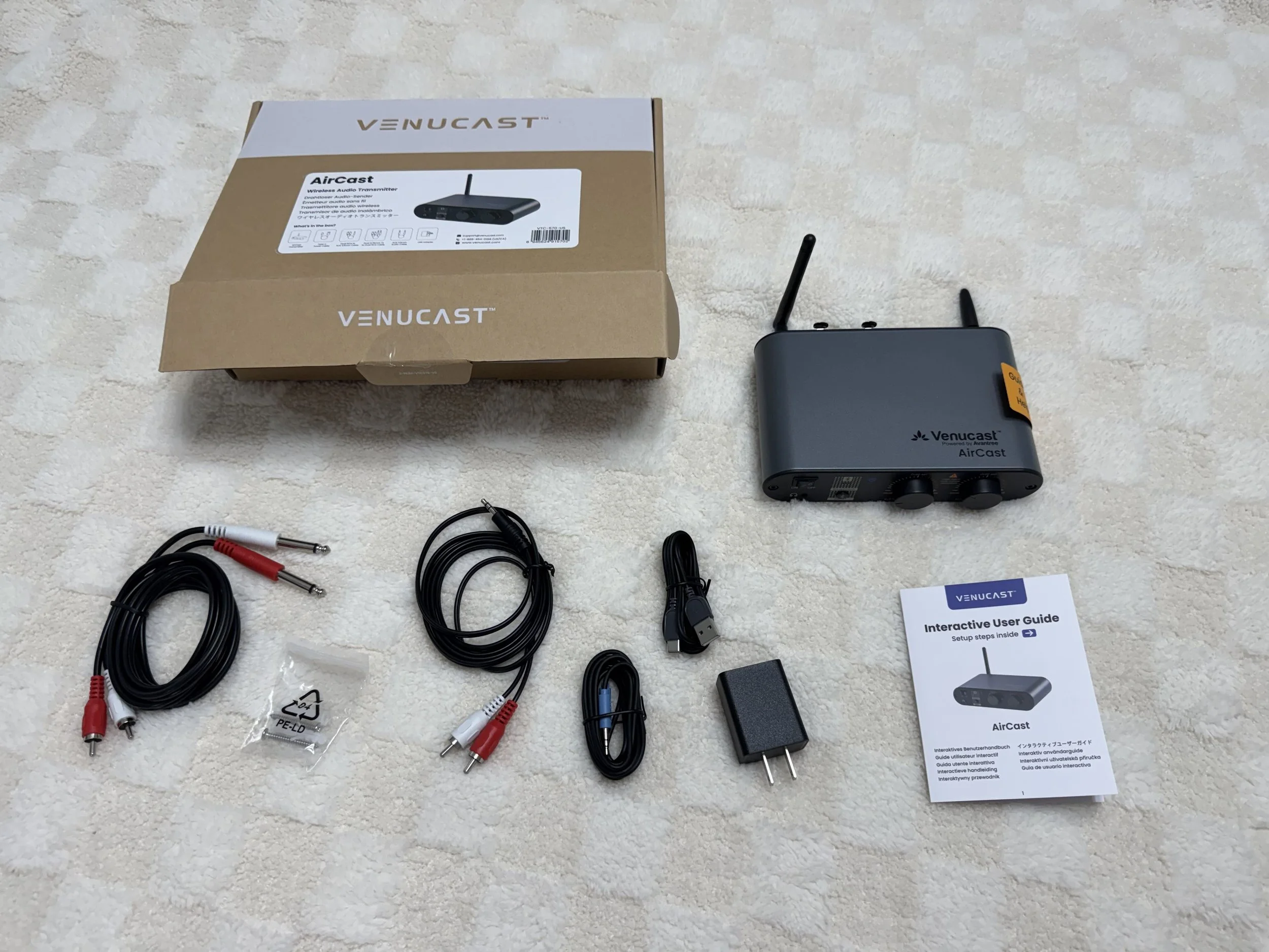 Unboxed Venucast AirCast wireless audio transmitter and accessories on a beige carpet, including cables, power adapter, user guide, and packaging box.