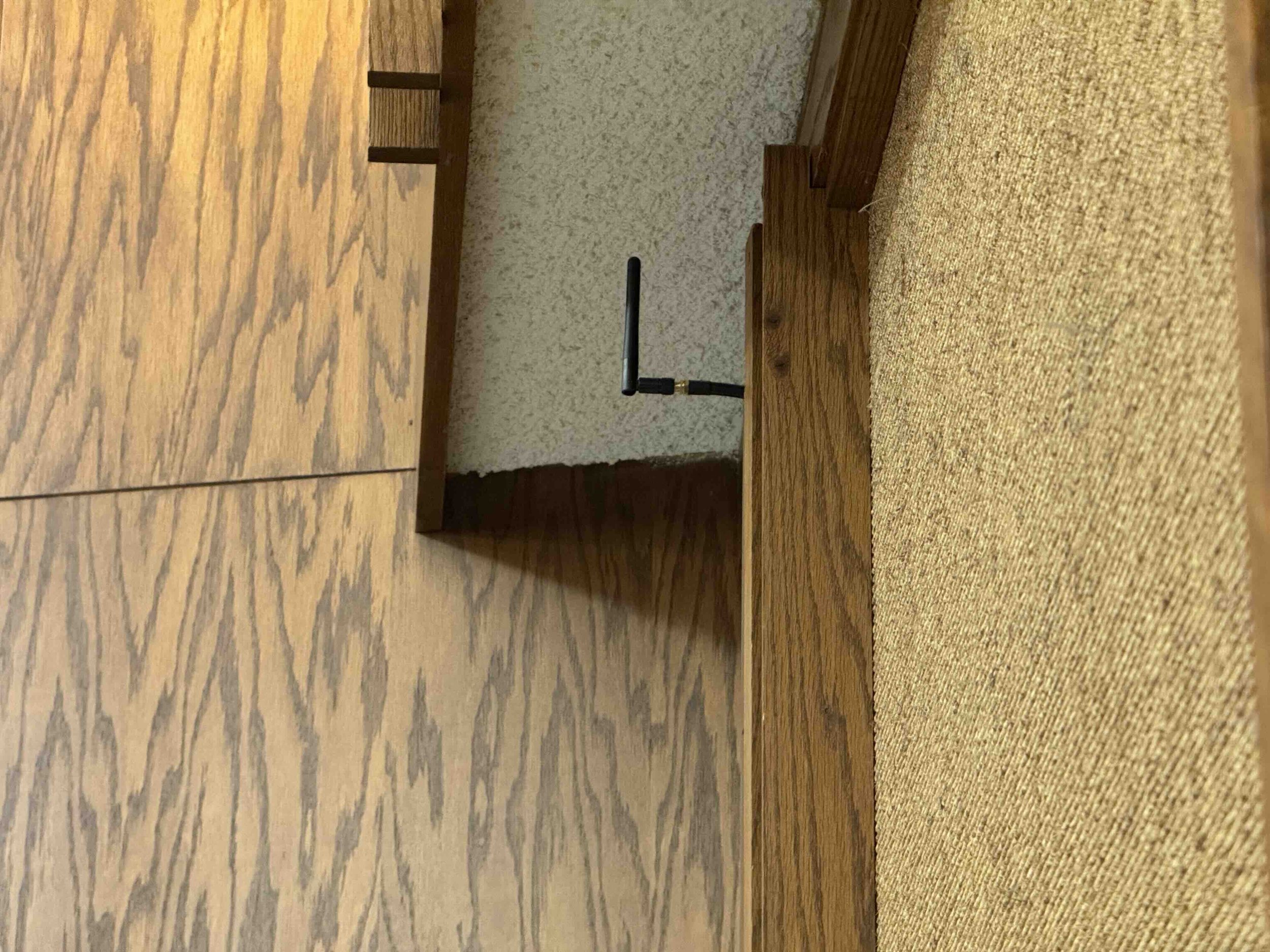 Close-up of a wooden table with a textured beige carpet on the right side. There is a small black object, possibly a clamp or antenna, attached to the edge of the table. The background shows a section of a wooden cabinet or panel and textured wall co
