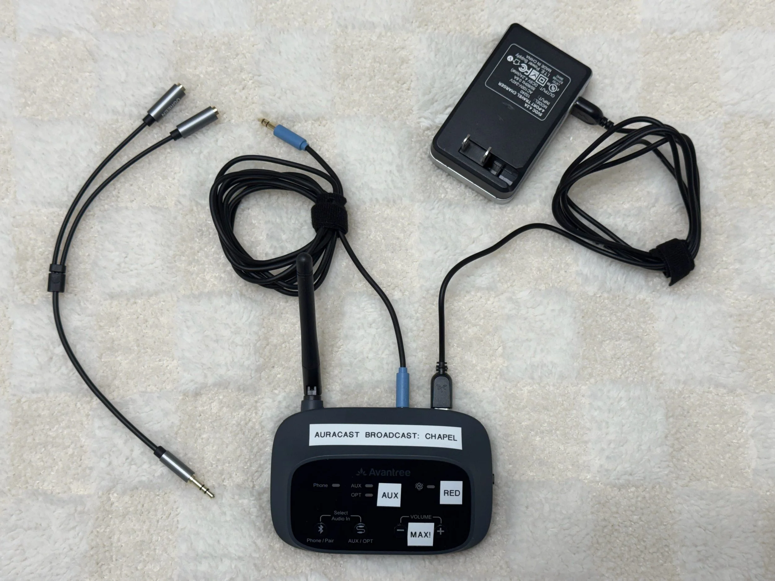 A small black electronic device with a display and labeled buttons, connected to several cables, with a black power adapter nearby on a beige surface.
