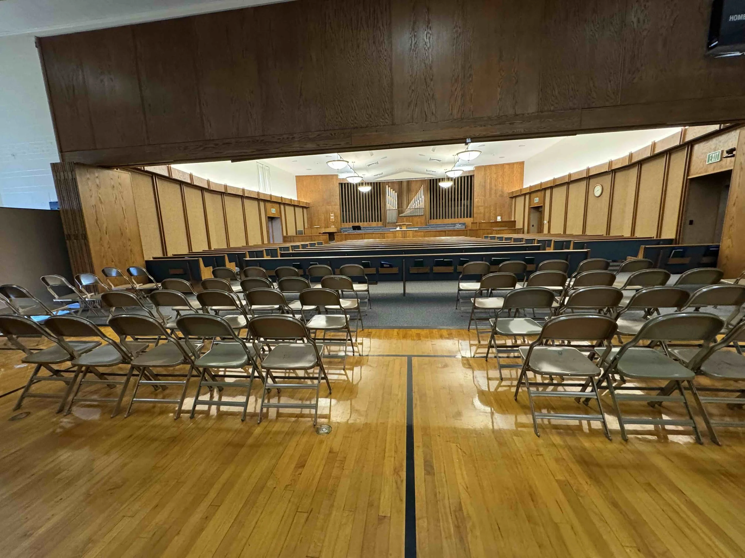 Empty church or auditorium with rows of chairs facing a raised stage area with organ pipes and light fixtures.