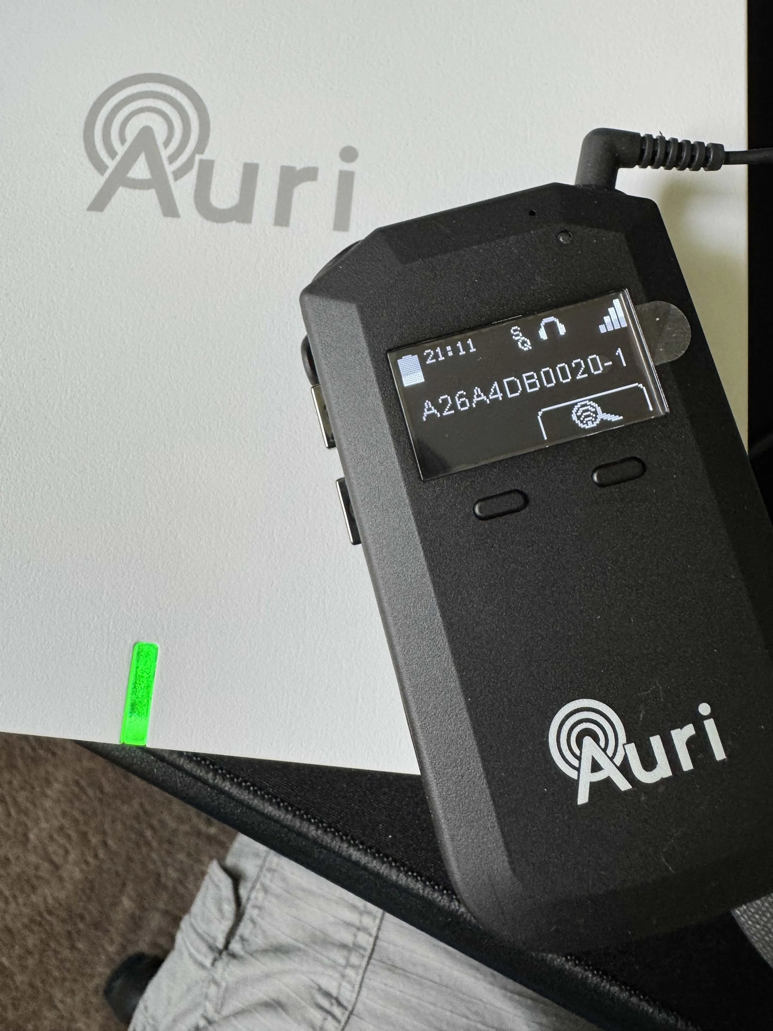A digital audio recorder on top of an Aura branded speaker or device. The recorder shows a time of 21:11 and has various icons and markings on its screen. In the background, there is a white wall with the Aura logo visible.