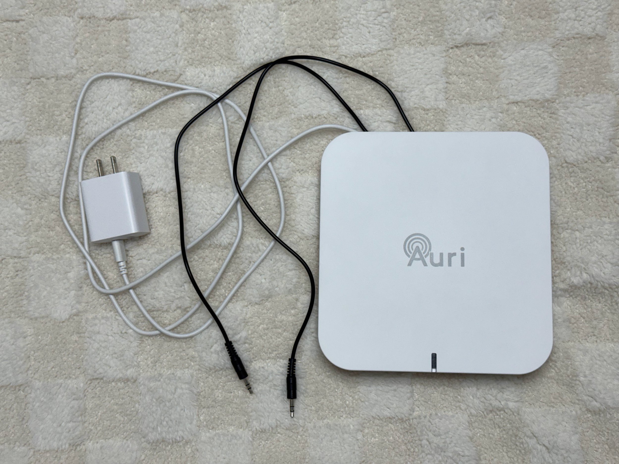 A white electronic device labeled 'Auri' with a wireless charging symbol, connected to a white power adapter and multiple black and white cables with audio jack connectors, all placed on a light-colored carpet.