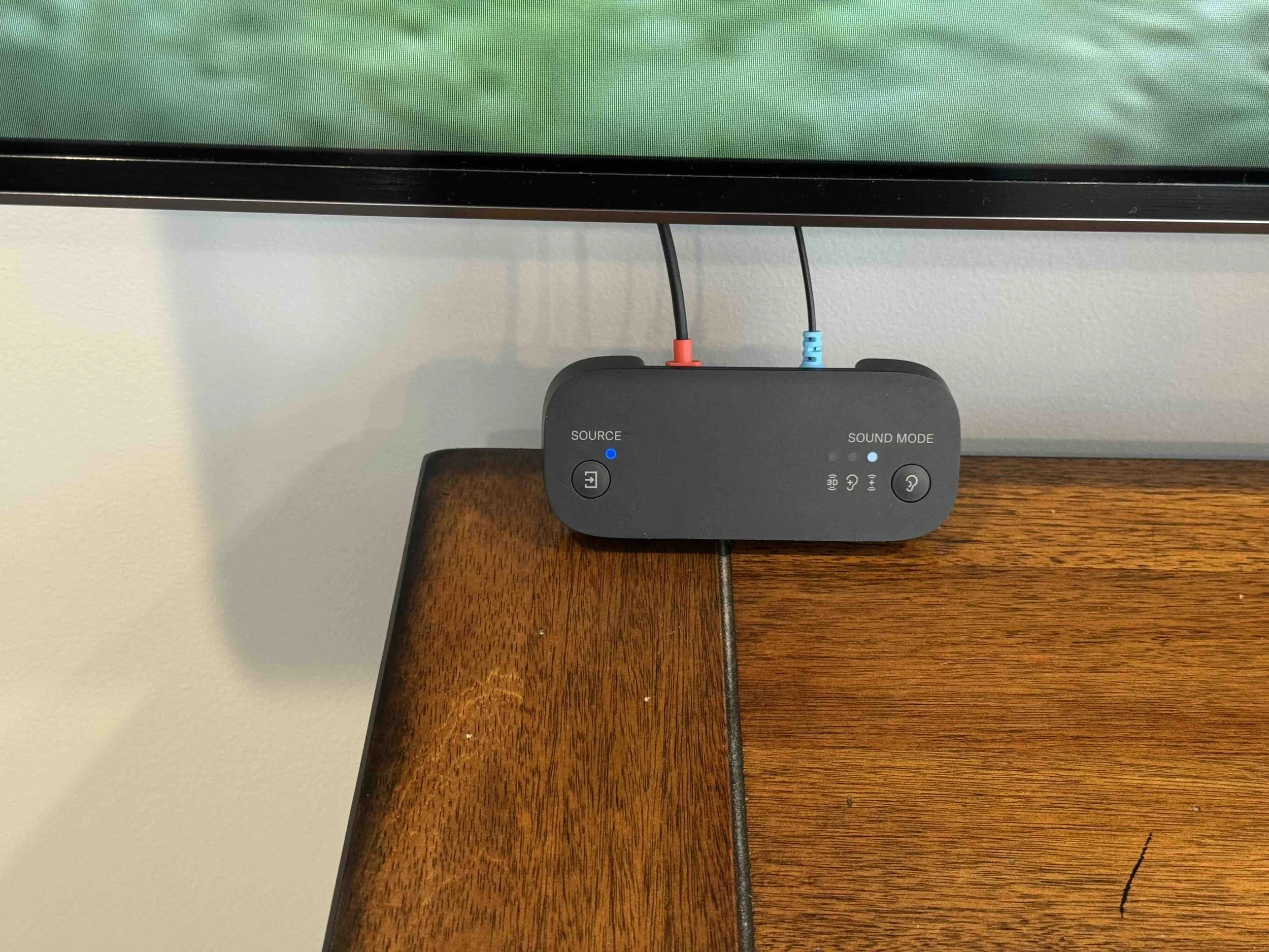 Close-up of a computer monitor on a wooden desk, with a black electronic device connected with red and blue wires visible below the monitor.