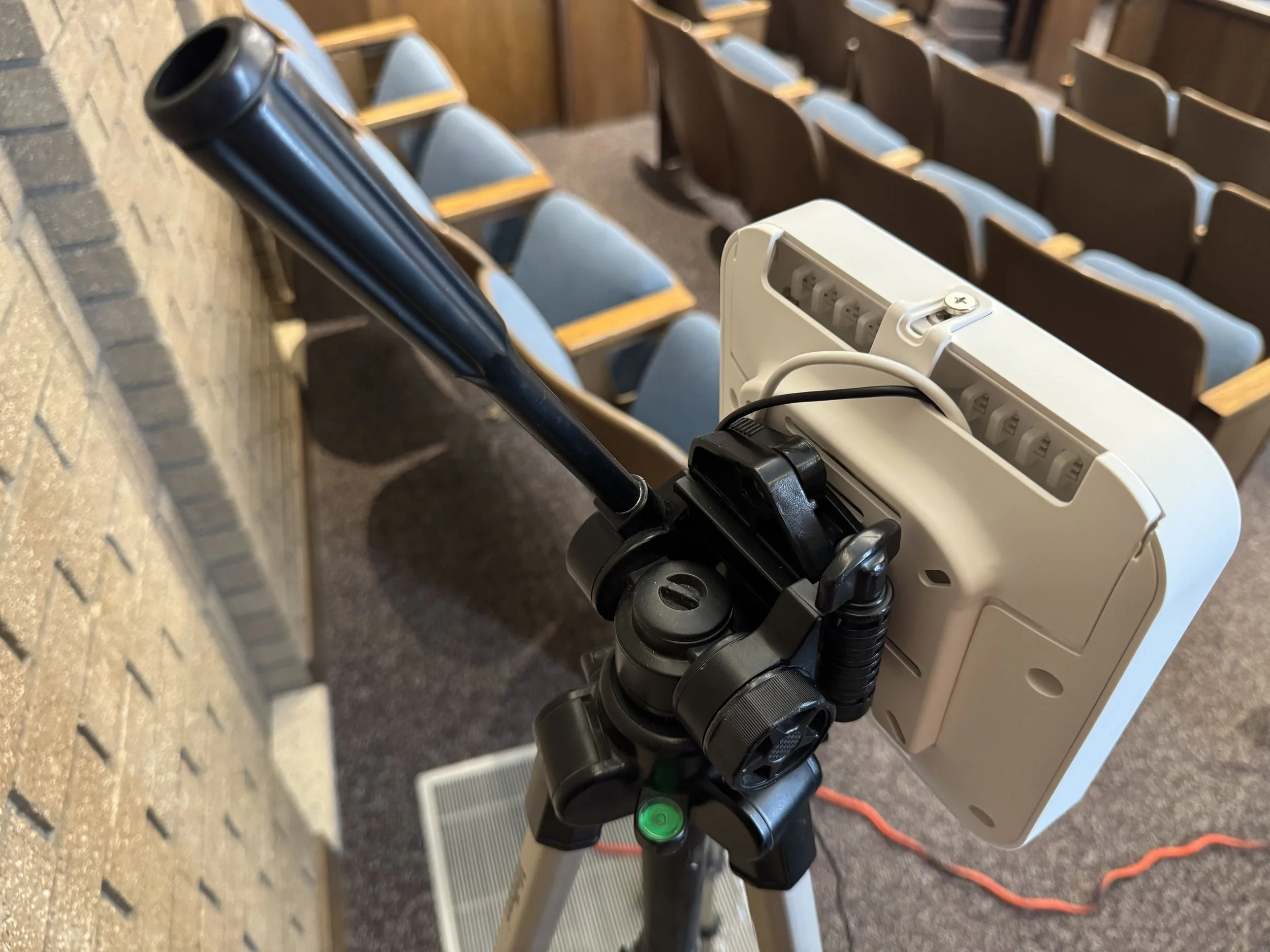 An electric wheelchair with a charging cord plugged into the back, positioned next to a wall with chairs in the background.