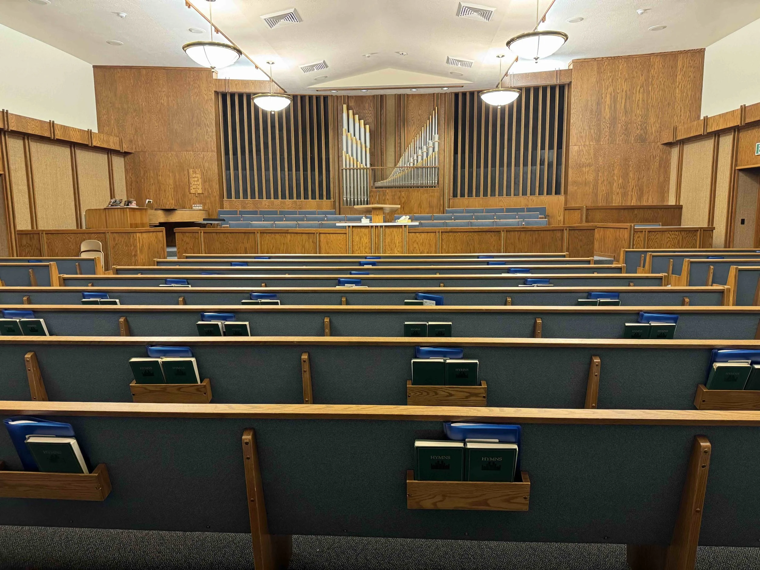 Interior of a church or chapel with rows of pews, a stage with organ pipes, and wooden paneling on the walls.