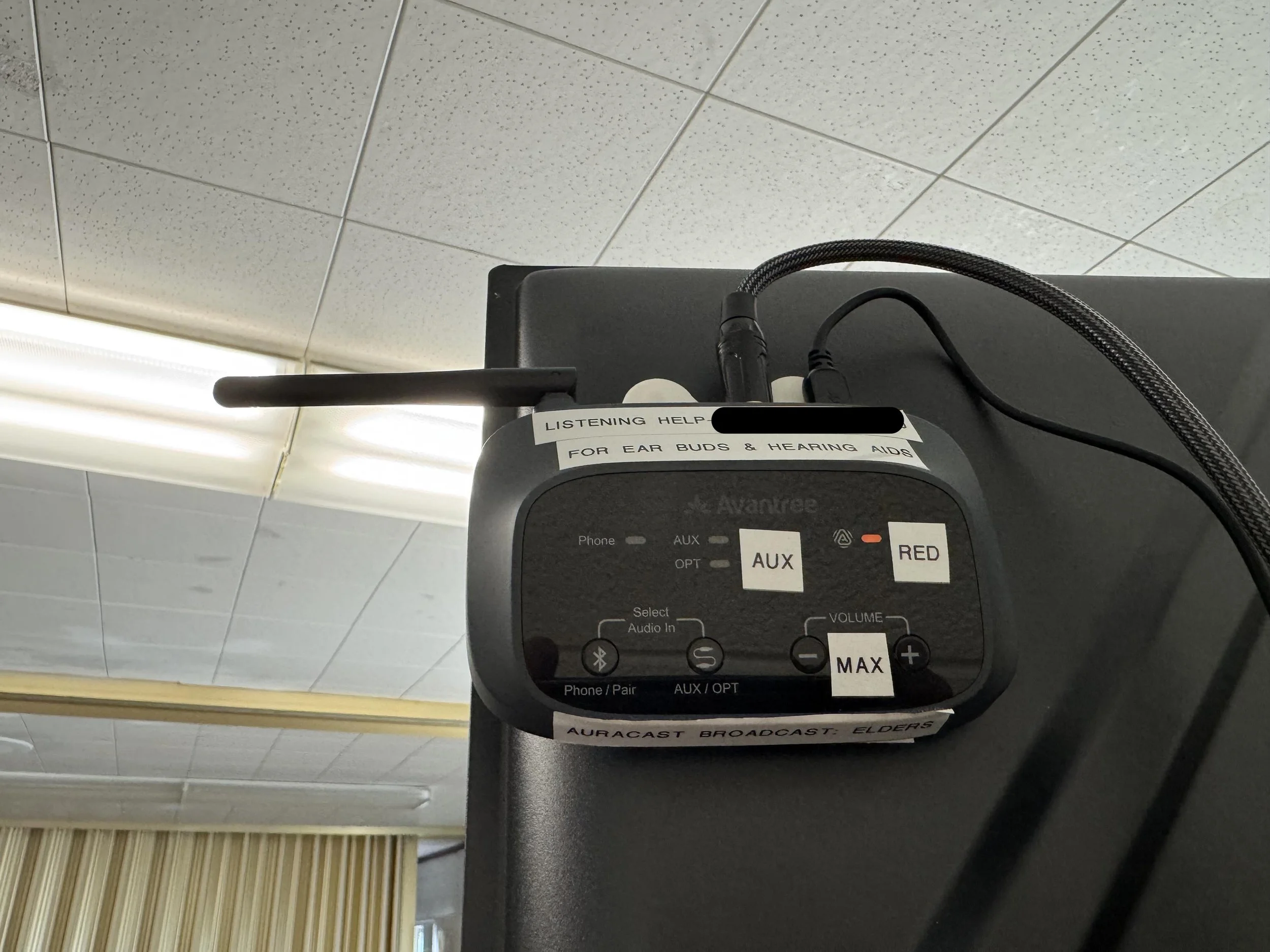 Close-up of a medical device with wires and labels, including a sticker that reads 'Listening Help for Ear Buds & Hearing Aids,' set against a tiled ceiling background.