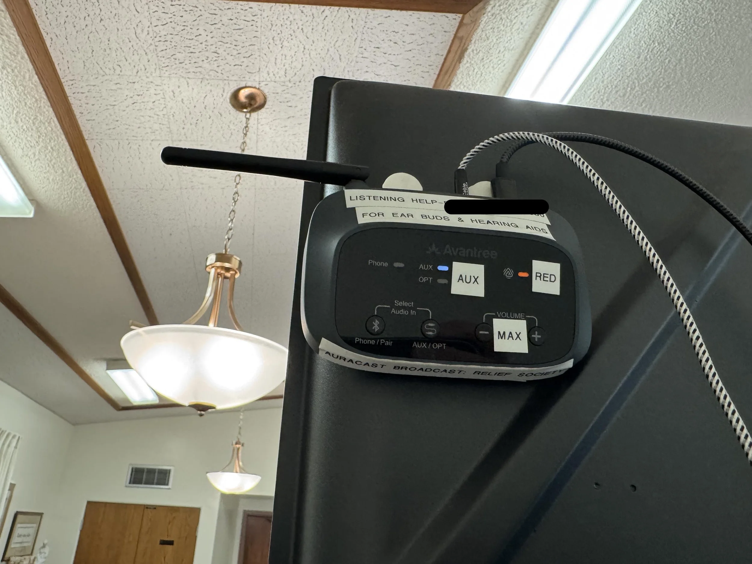 Close-up of a hearing aid device with a label that reads 'Listening Help' for ear buds and hearing aids, positioned on a black tray with visible cables and controls, in a room with ceiling lights and a window.