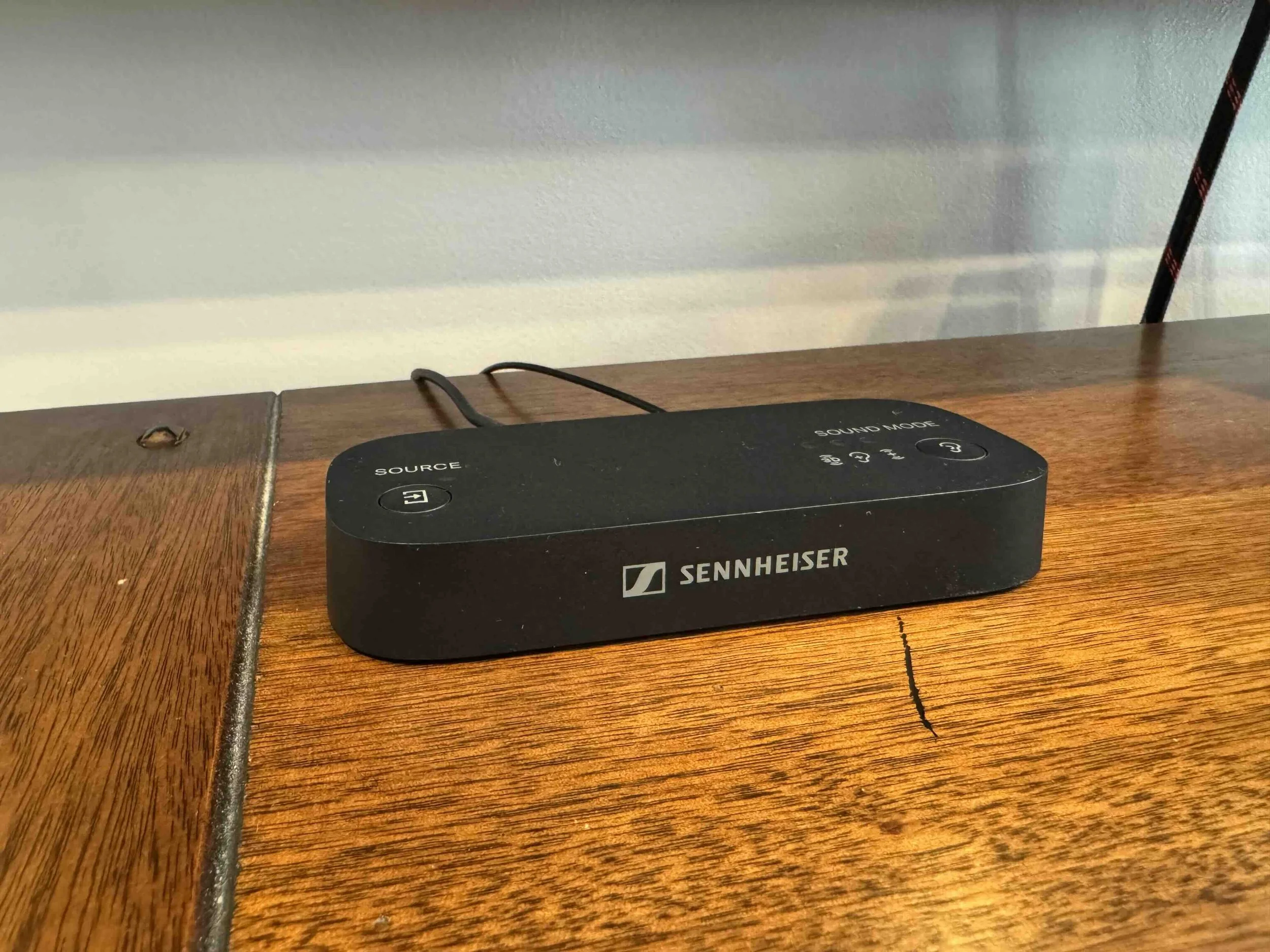 A black Sennheiser wireless receiver or transmitter with labeled buttons for source and sound mode, placed on a wooden surface with a visible screw, against a wall with a faint shadow.