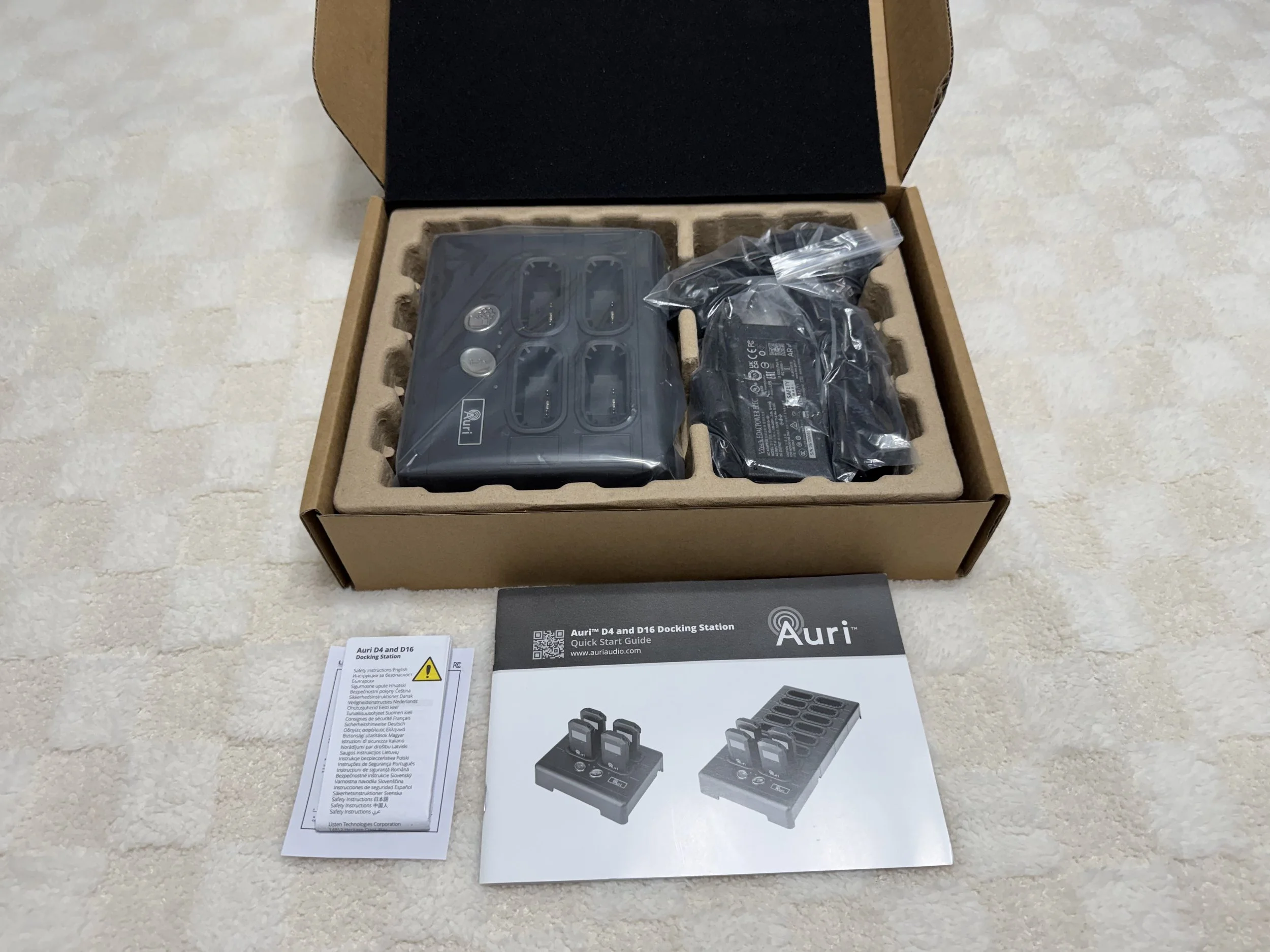 Open cardboard box containing black electronic device parts and accessories, including a controller and power adapter, with instruction manuals and product information sheets placed in front of the box.