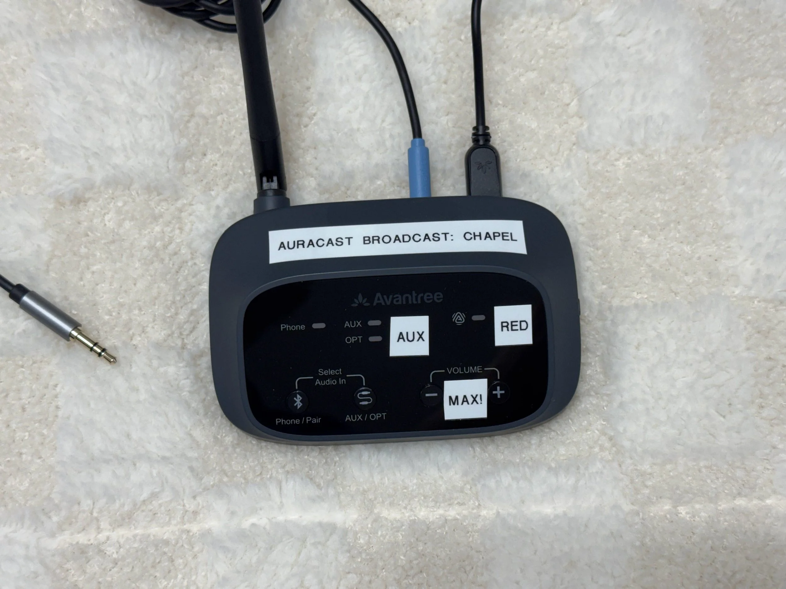 A handheld electronic device labeled 'Avantree' with various black, white, and red buttons and labels. The device has three cables plugged into it: a black cable, a blue cable, and a black cable with a symbol of a fly. An audio jack connector lies on