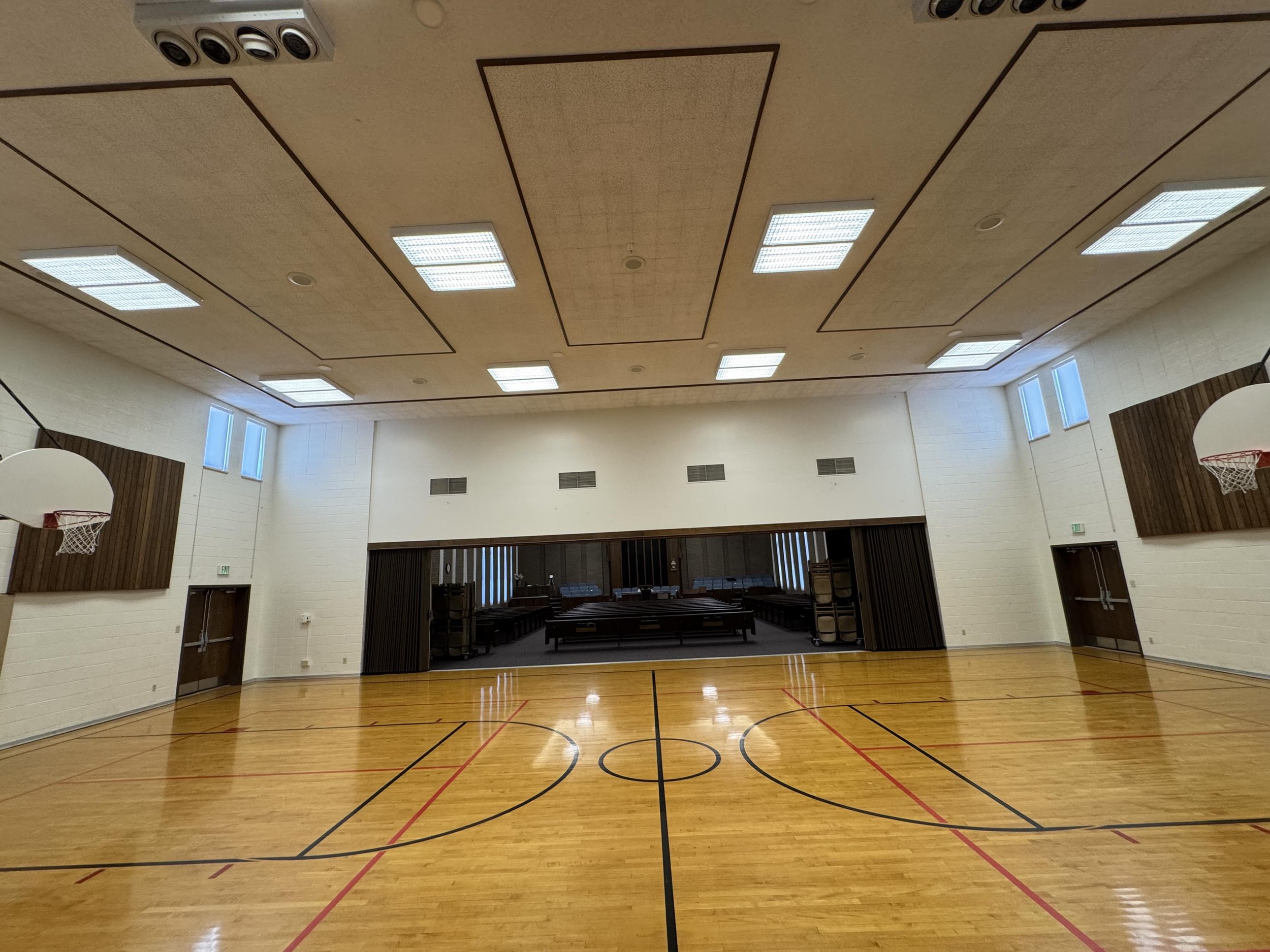Indoor gymnasium with a polished wooden floor, basketball hoops, and high windows, illuminated by ceiling lights.