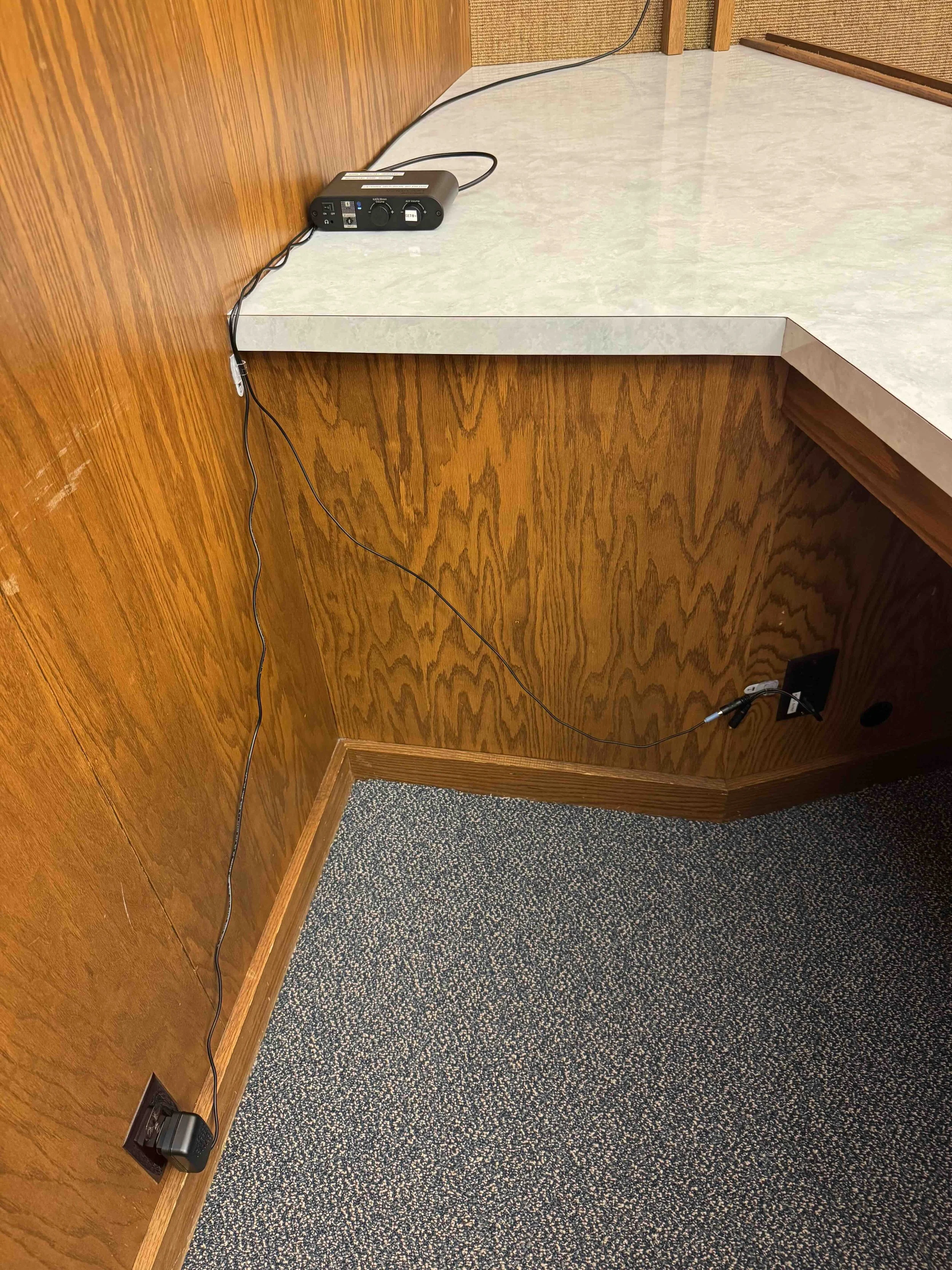 Corner of a room with wood-paneled walls, a gray countertop, power outlets, a black power strip, and a carpeted floor.
