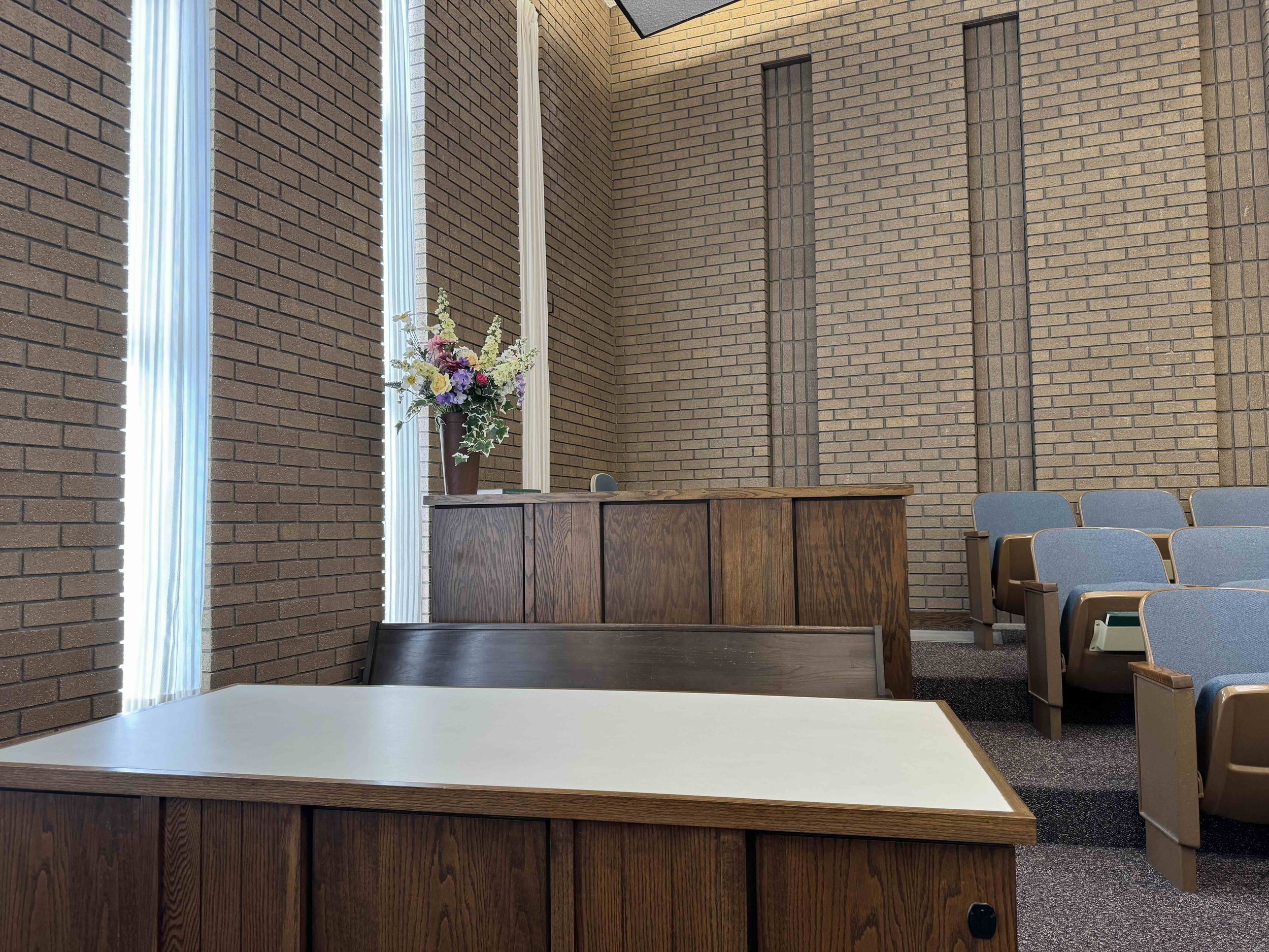Interior of a room with brick walls, a wooden desk, a flower arrangement in a brown vase on the desk, blue cushioned chairs, and a bright vertical window with blinds.