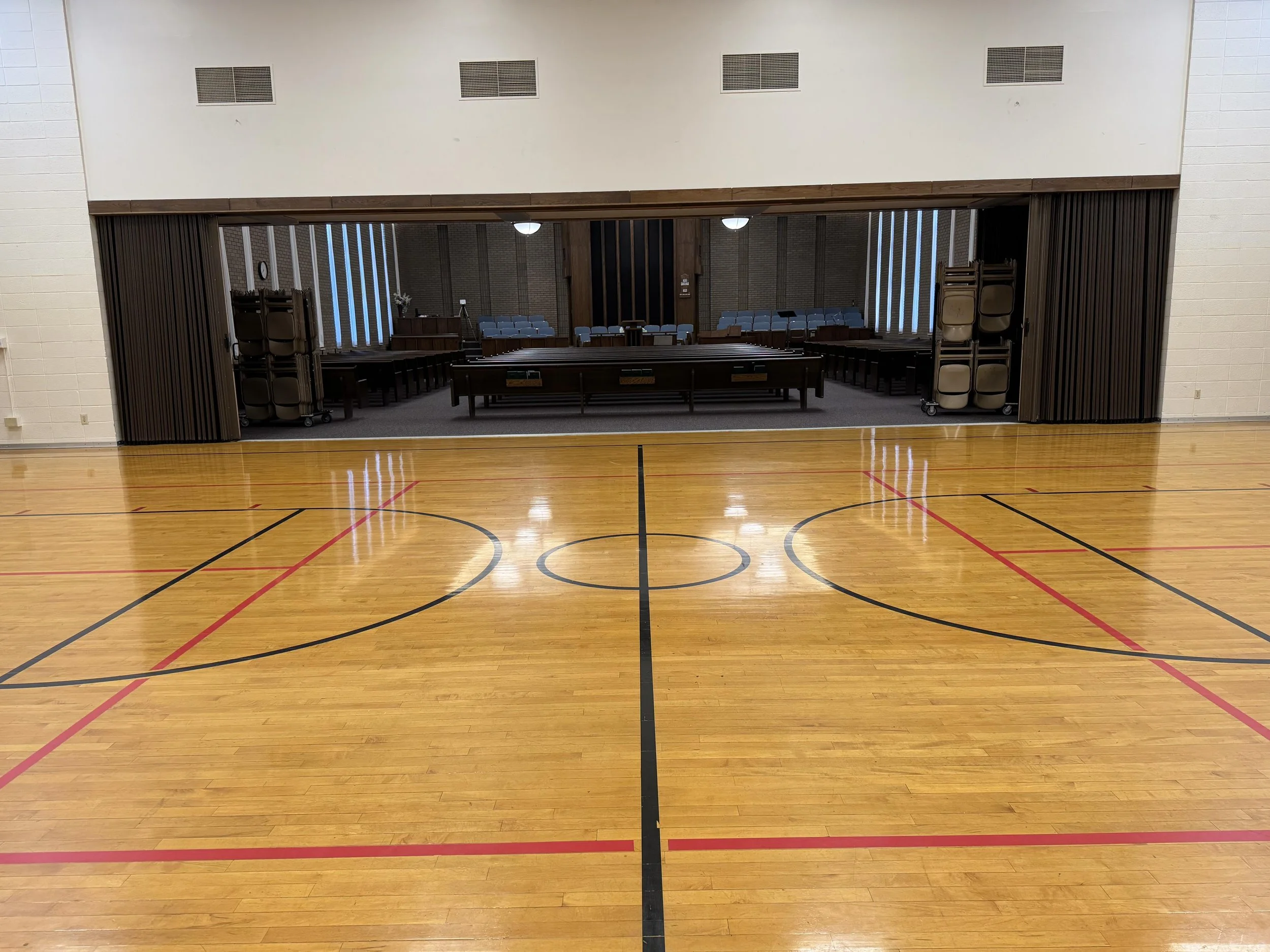 Empty gymnasium with a stage visible through large open doors, lined with chairs, and a wooden floor with black and red court markings.