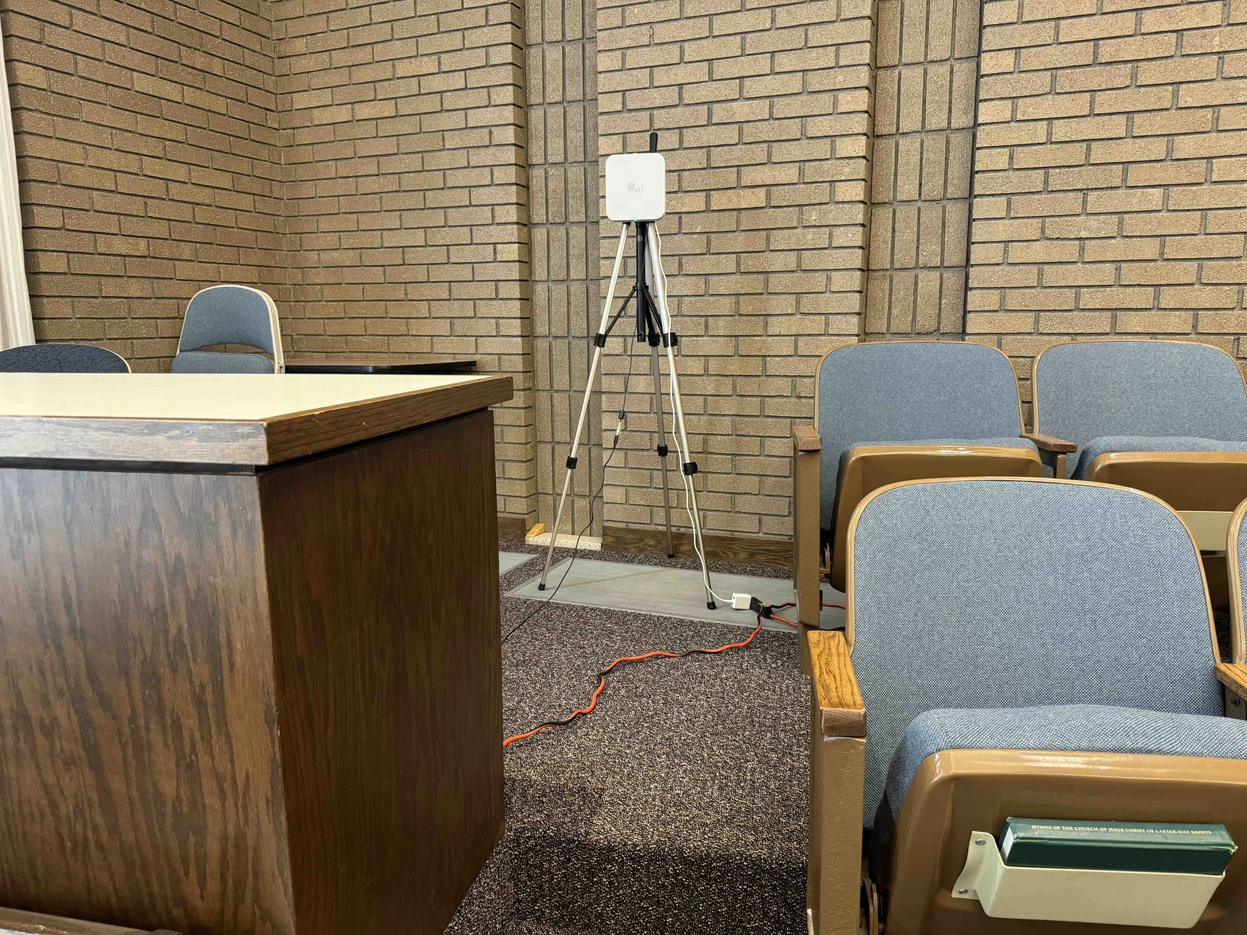 Interior of a meeting room with blue cushioned chairs, a wooden pulpit, a brick wall, and electronic equipment on a tripod, including a white device labeled Auri and an orange extension cord on the carpeted floor.