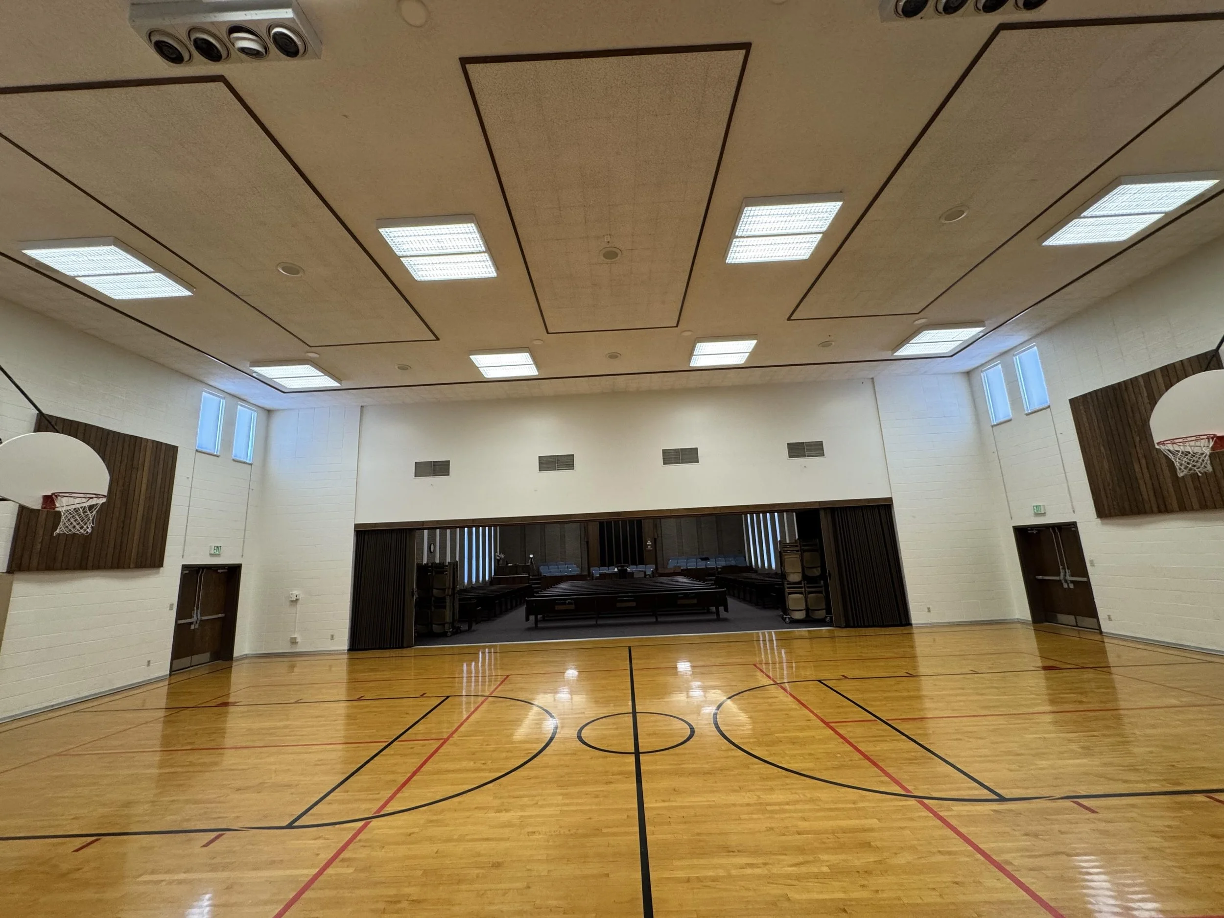 An empty indoor gymnasium with a basketball hoop, wooden flooring, and high windows.