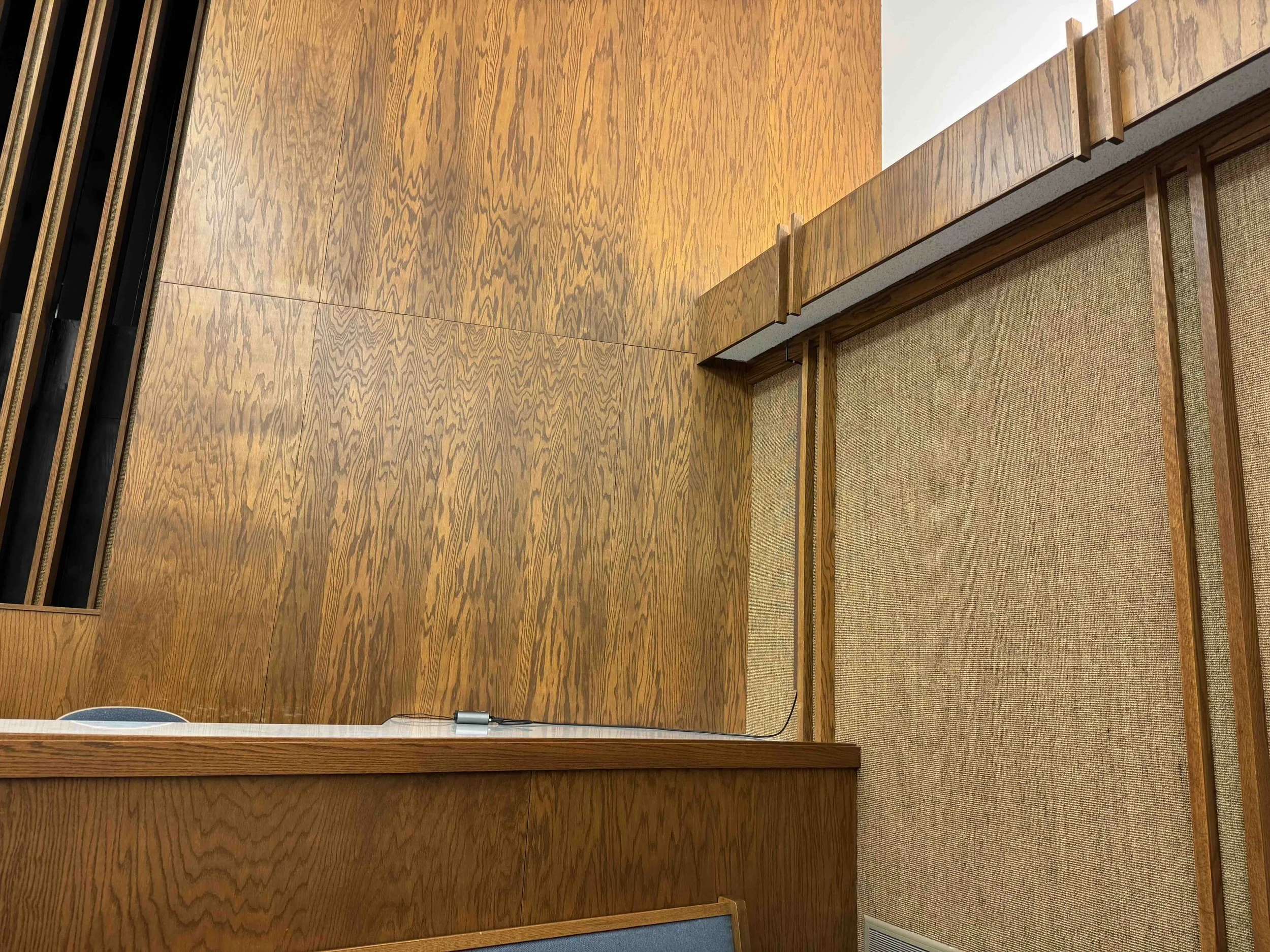 Wood-paneled wall with a black grate on the left and a small table or ledge at the bottom with a camera and a cable.
