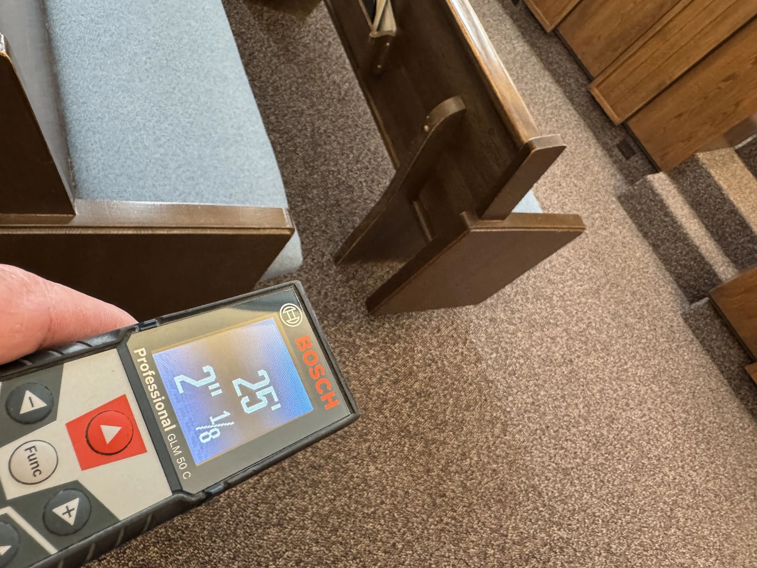 A handheld electronic device displaying a digital glucometer screen with a reading of 125 mg/dL, surrounded by a carpeted floor, wooden furniture, and a blue upholstered piece of furniture in the background.