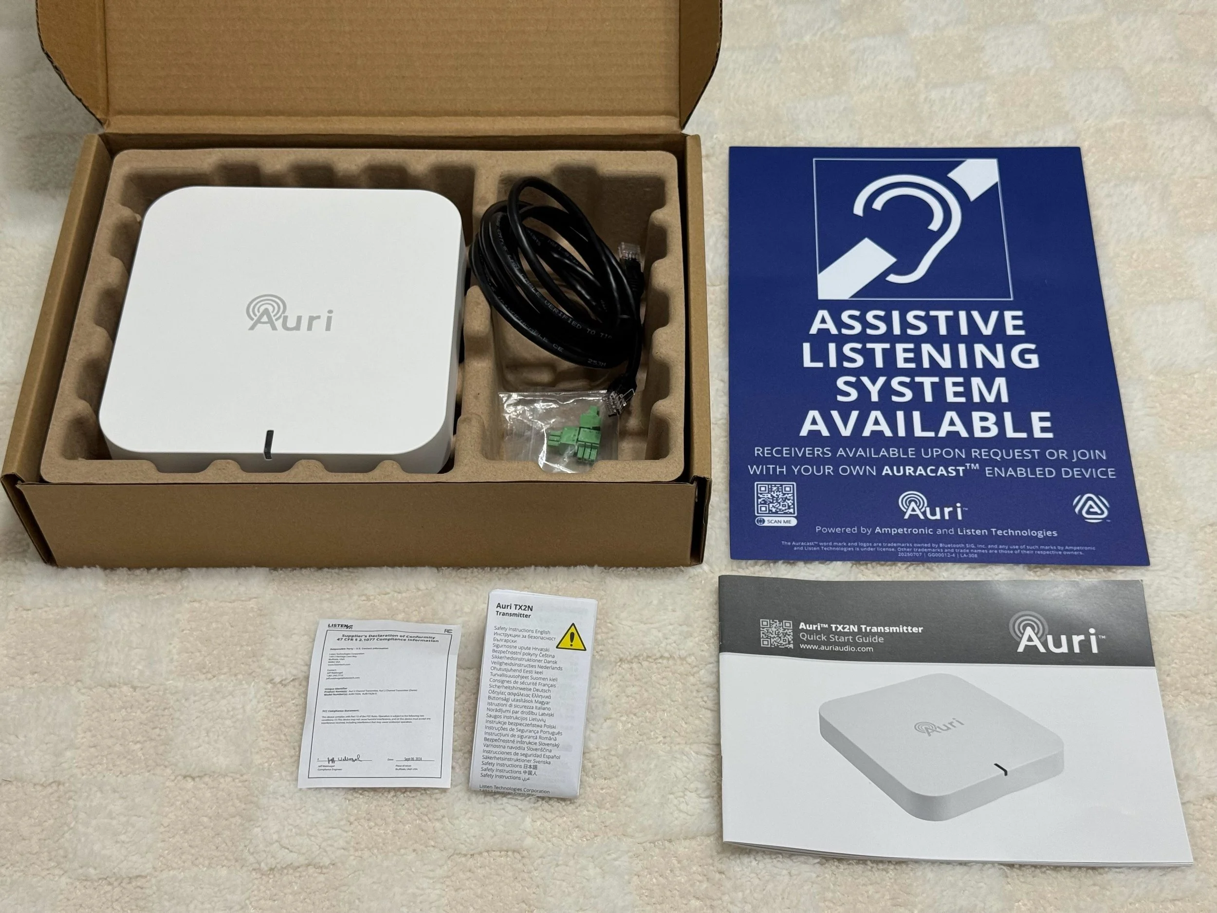 Open cardboard box containing a white Auri device, cables, and small electronic components. Beside the box is a blue sign indicating assistive listening system available, and instructional pamphlets for the Auri TX2N transmitter and quick start guide