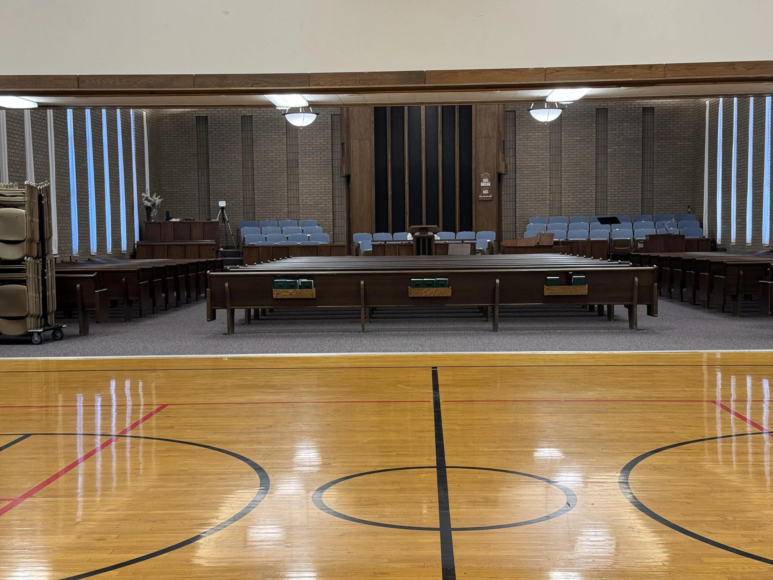 An indoor gymnasium with wooden flooring, brick walls, and blue cushioned chairs arranged in rows at the front. A podium or altar with flowers, a table with a tripod, and a clipboard are visible at the back. The ceiling has large light fixtures, and 