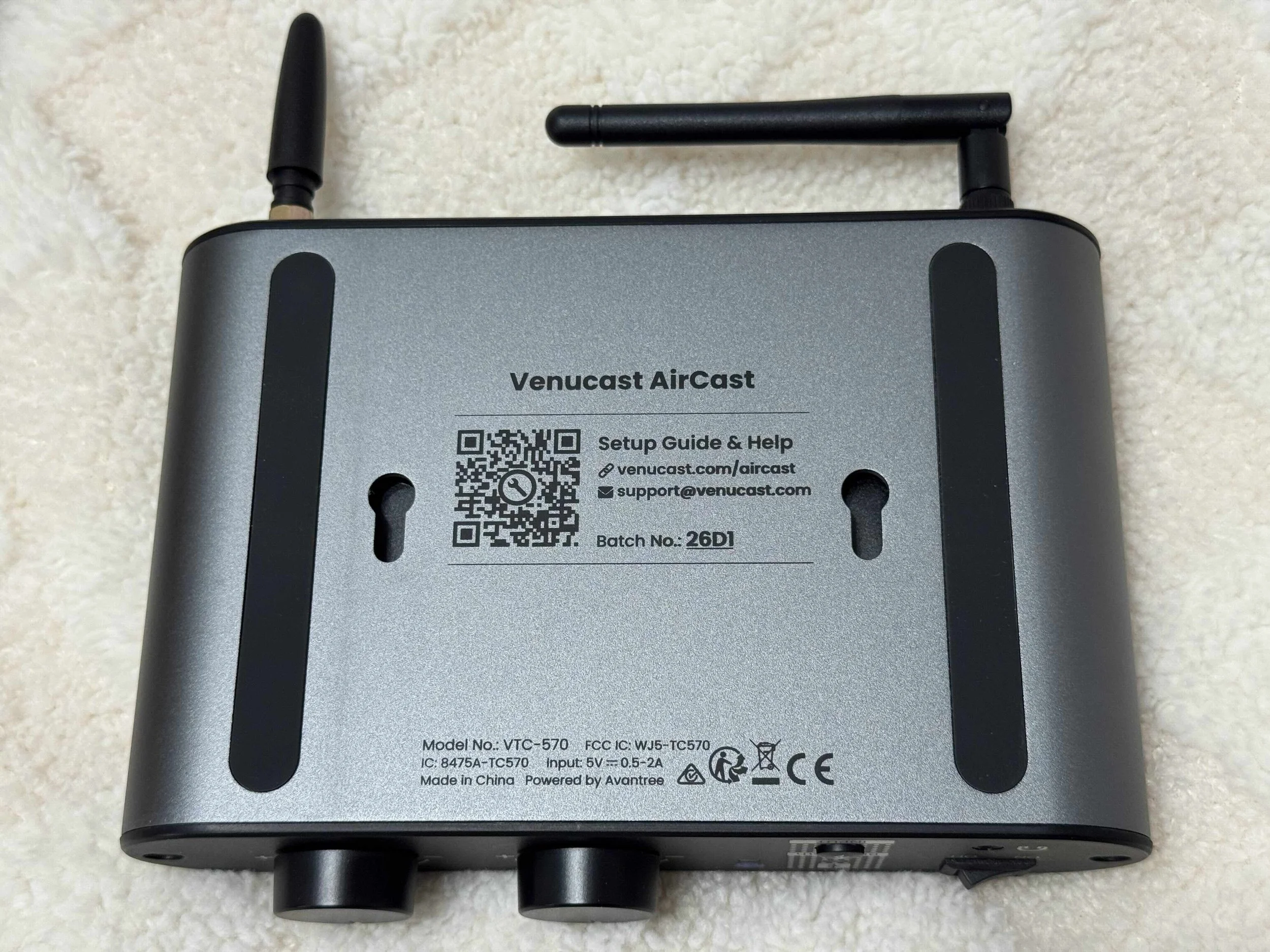 A Venucast AirCast device with antennas, QR code, model number VTC-570, and various symbols and text on its label, placed on a light-colored textured surface.