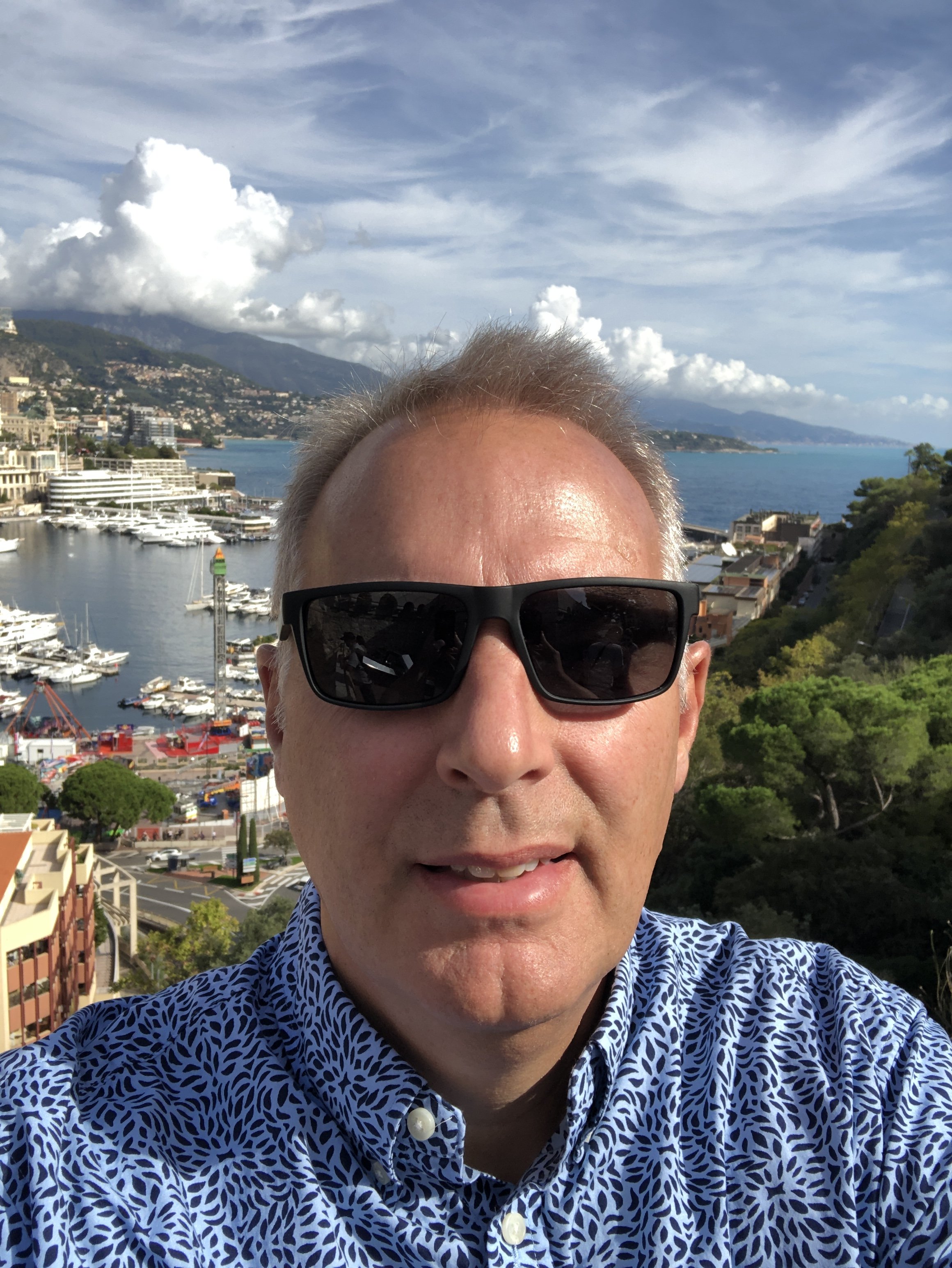A man wearing sunglasses and a patterned shirt taking a selfie with a coastal city and harbor in the background, featuring boats and mountains under a partly cloudy sky.