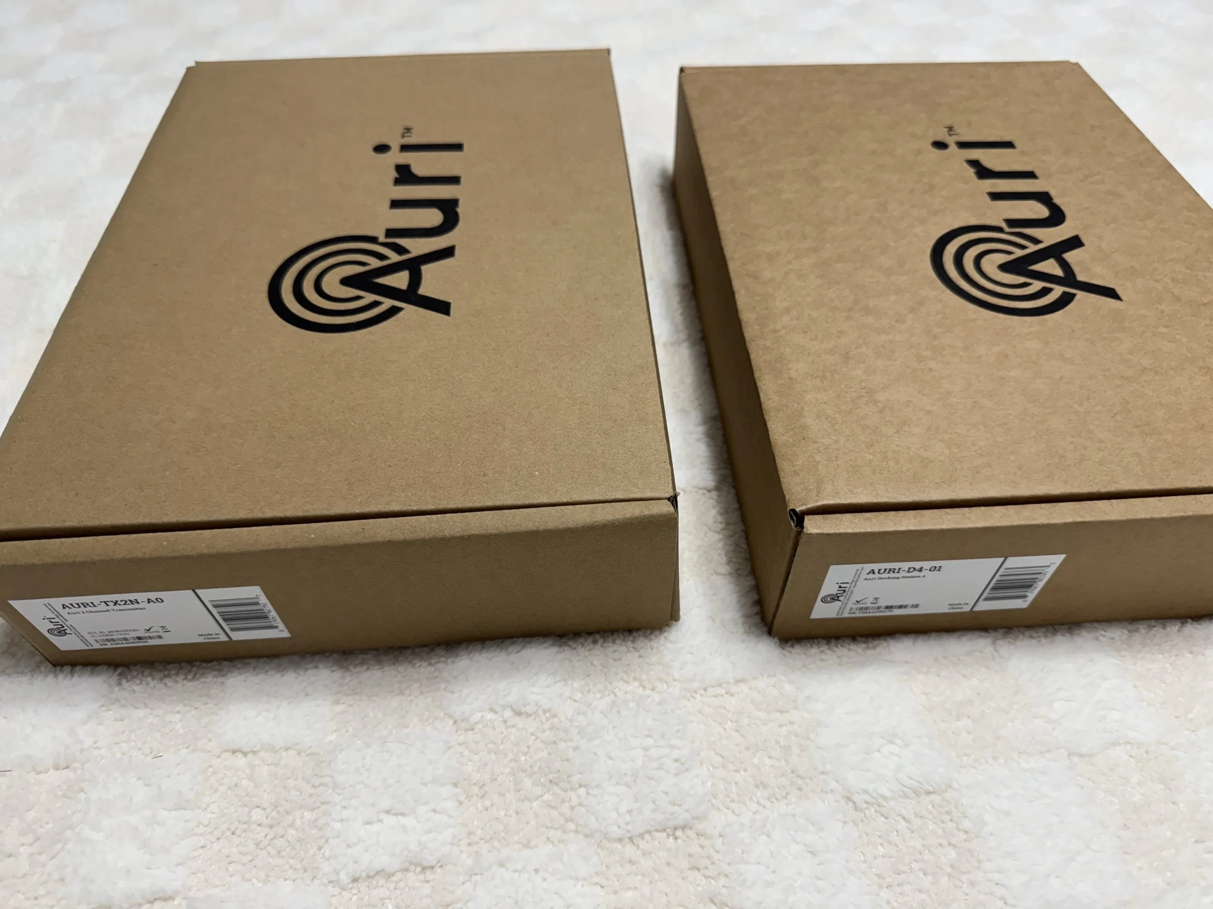 Two cardboard boxes placed side by side on a white carpeted surface, each with a label and a barcode at the bottom and the word 'AURI' printed in black on the top.
