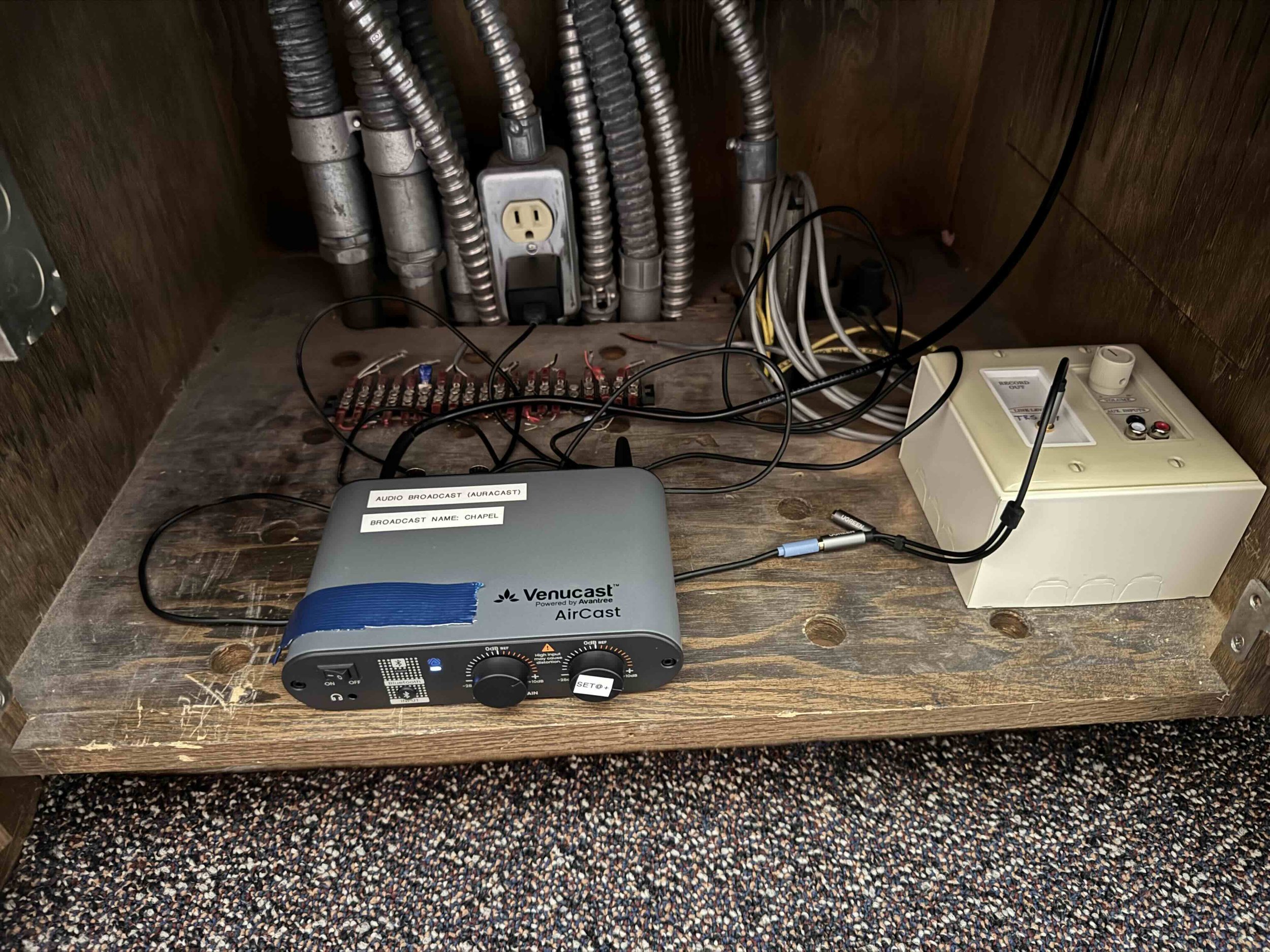 A wooden table with electrical equipment, including a gray Venucast audio broadcast device, a white power strip, and multiple wires and cables connected to the equipment, all set in a small enclosed space with wooden walls.