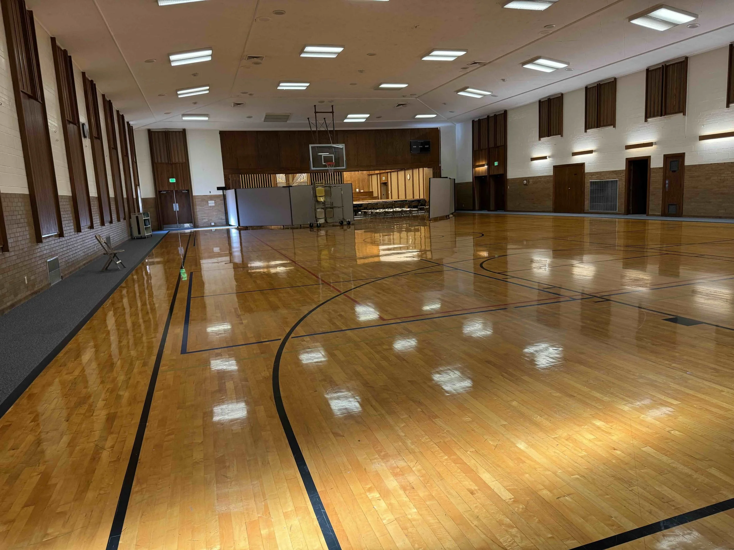 Empty indoor basketball court with polished wooden floor, basketball hoop, and chairs stacked in the background.