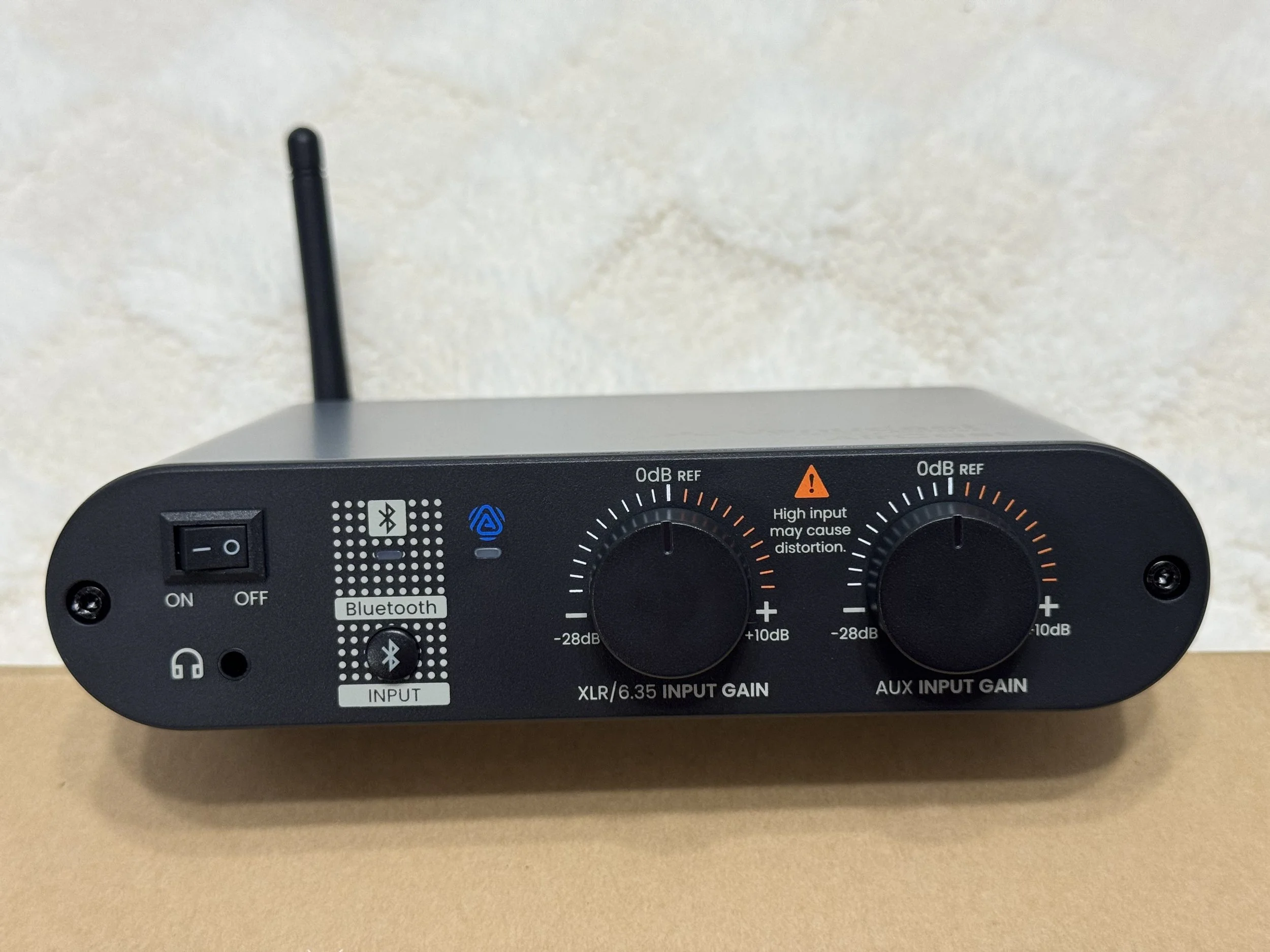 Close-up of a black audio interface device with various input controls, symbols for Bluetooth, and a power switch, placed on a beige surface.