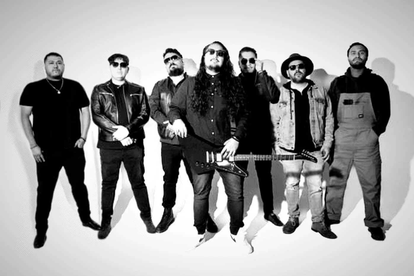 A group of eight men standing against a plain background, with one holding an electric guitar. The image is in black and white.