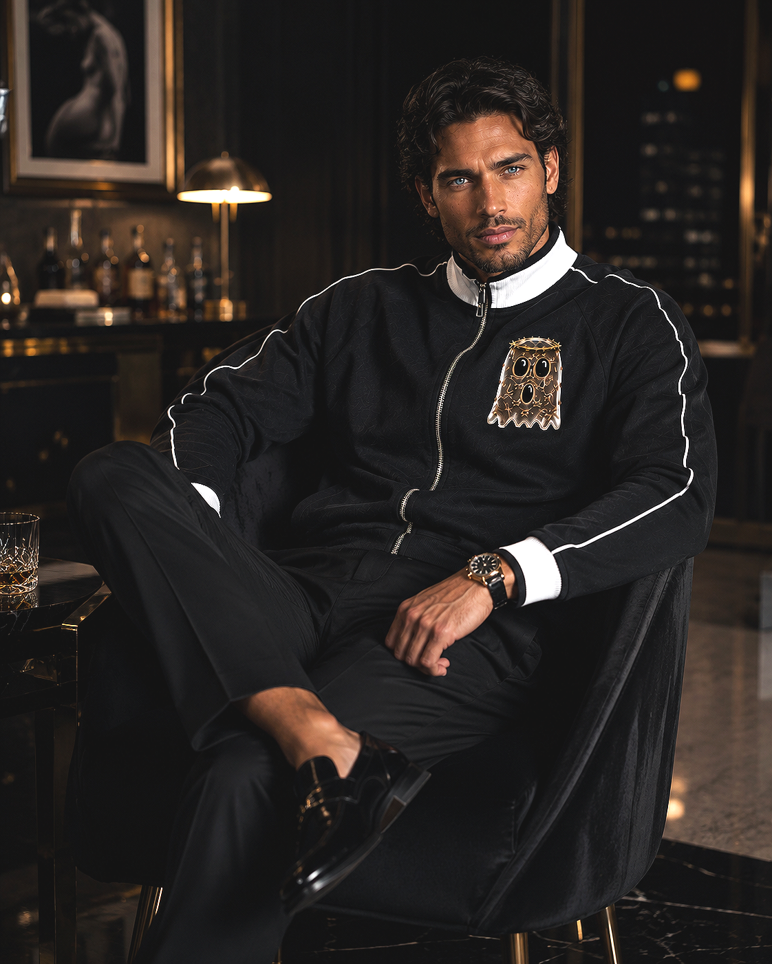 Stylish man sitting in a luxurious lounge, wearing a black jacket with a playful ghost patch, black pants, and shiny black shoes, with a glass of whiskey on the table beside him.