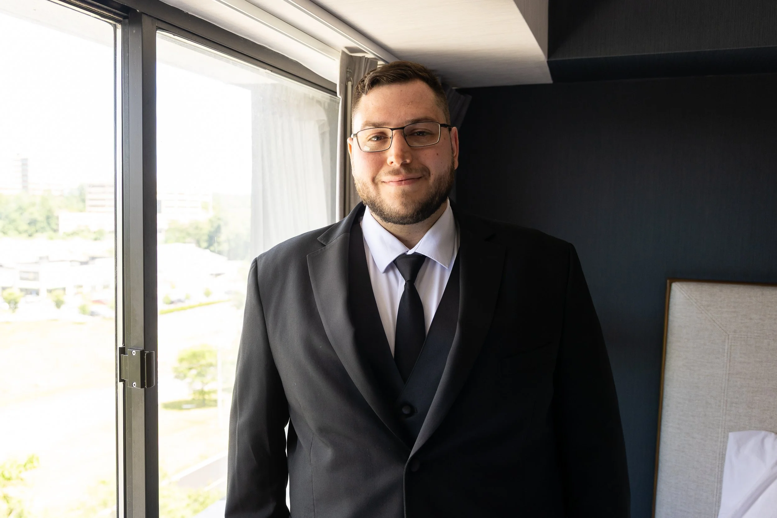 Man in a black suit, white shirt, and black tie smiling indoors near a window with cityscape view.