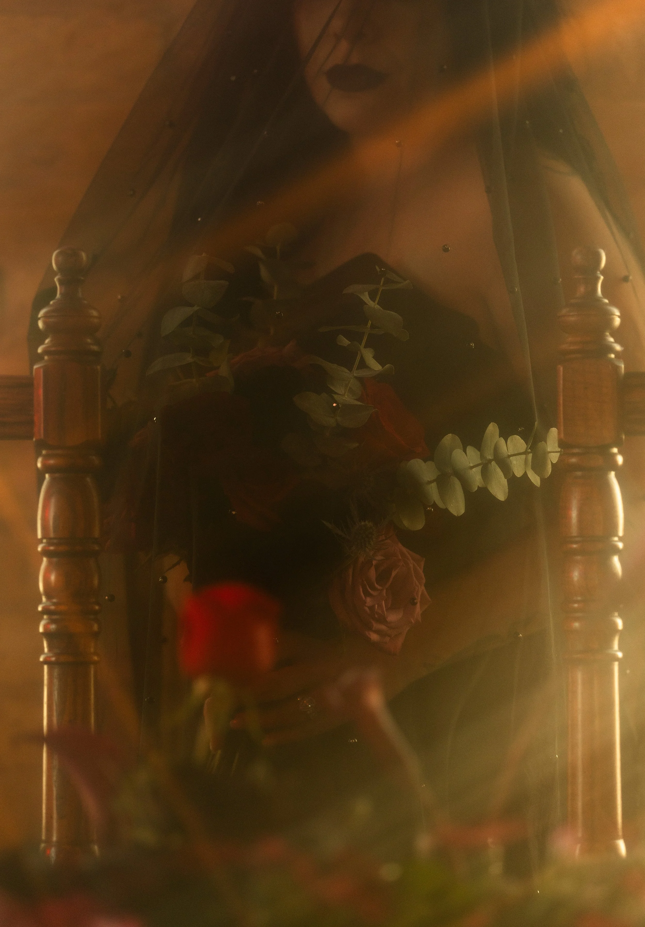 A reflection of a person taking a photo with their phone in a mirror on a dark wooden dresser. The dresser has turned posts and a decorative drawer handle. A cluster of eucalyptus leaves and a pink flower are visible on the dresser.
