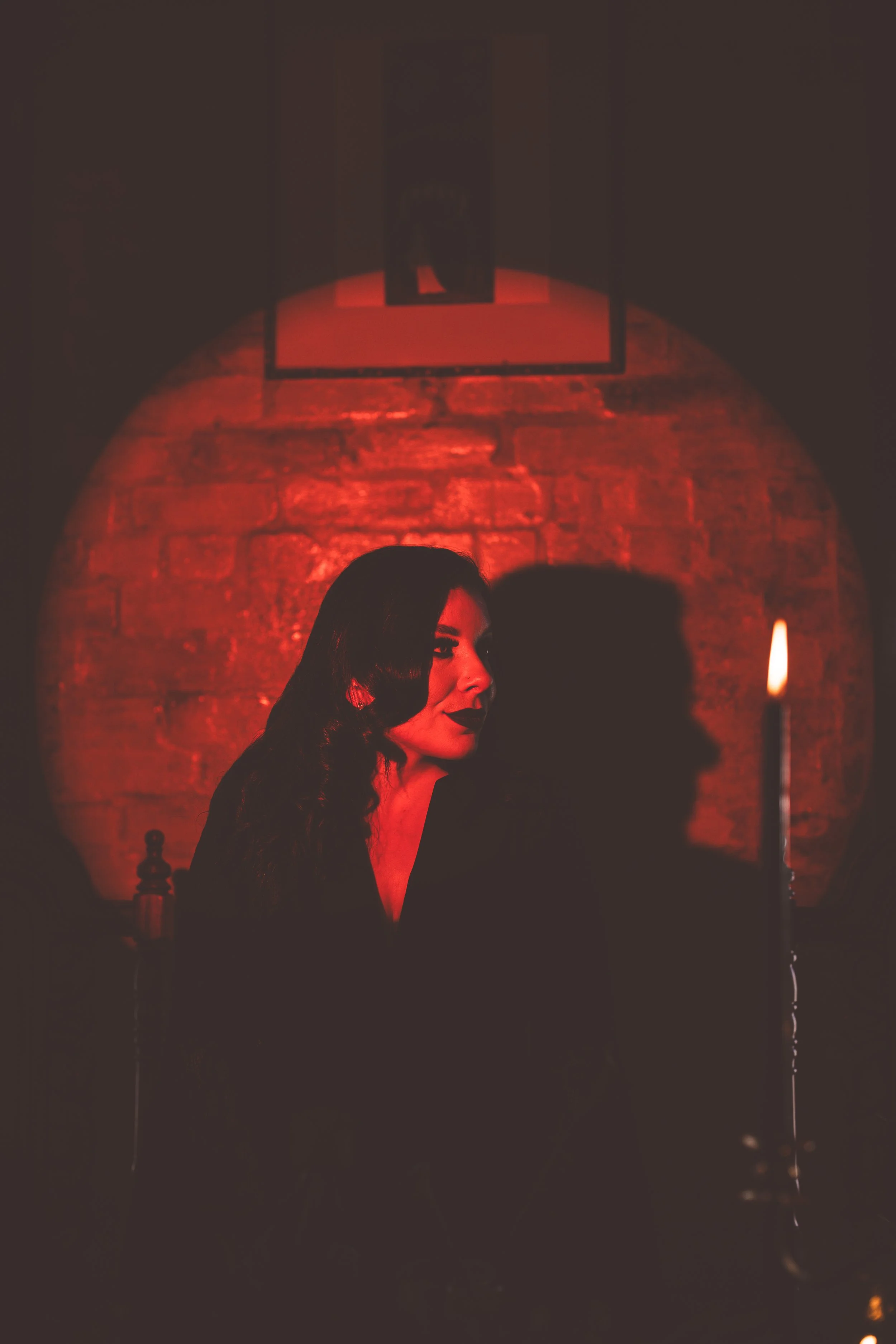 A woman with dark hair and makeup sitting in a dimly lit room with red lighting, a brick wall, and a framed picture hanging on the back wall.