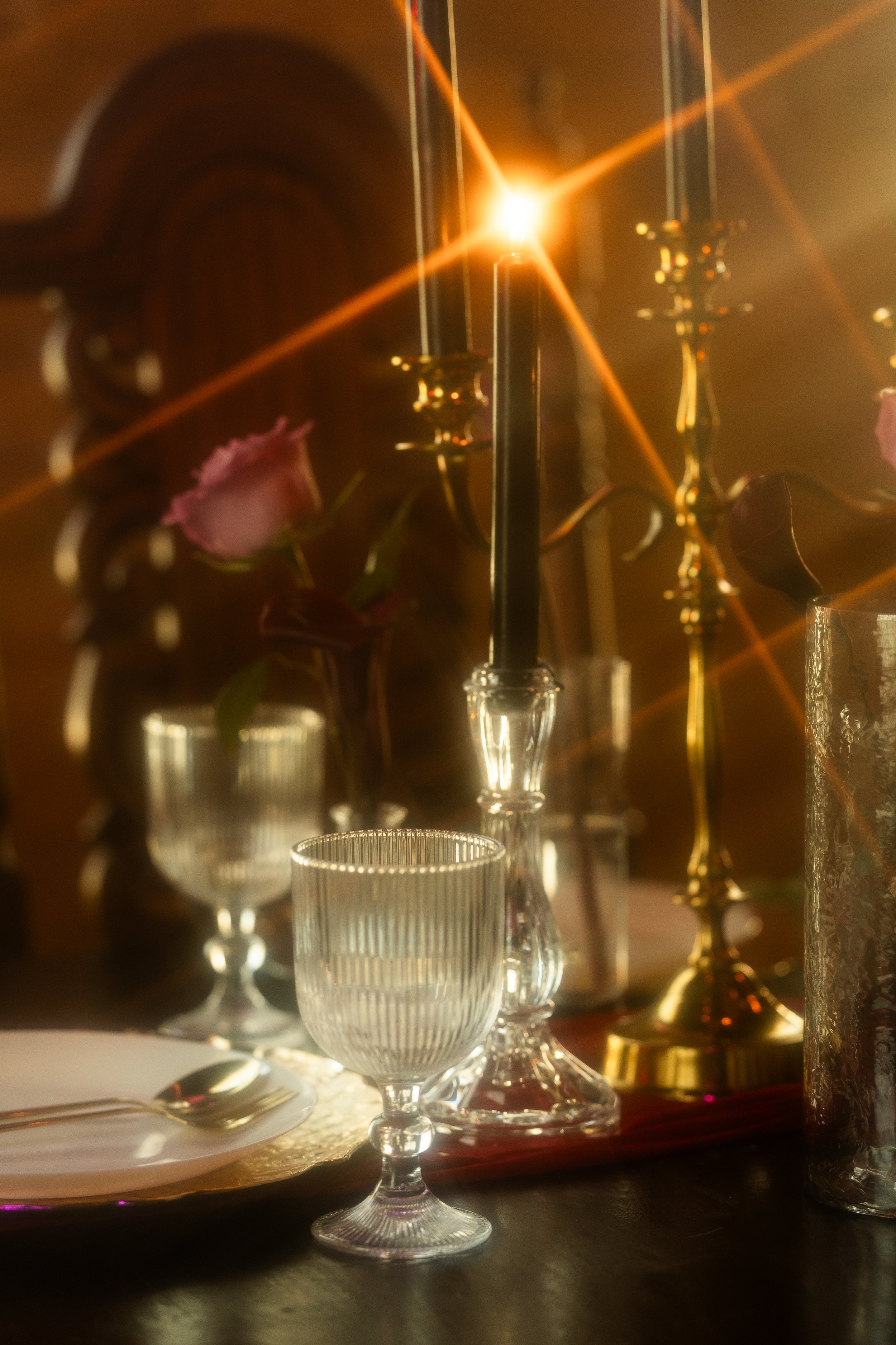Elegant table setting with wine glasses, a plate, cutlery, a pink flower, and candle holders, illuminated by warm lighting and a bright light source in the background.