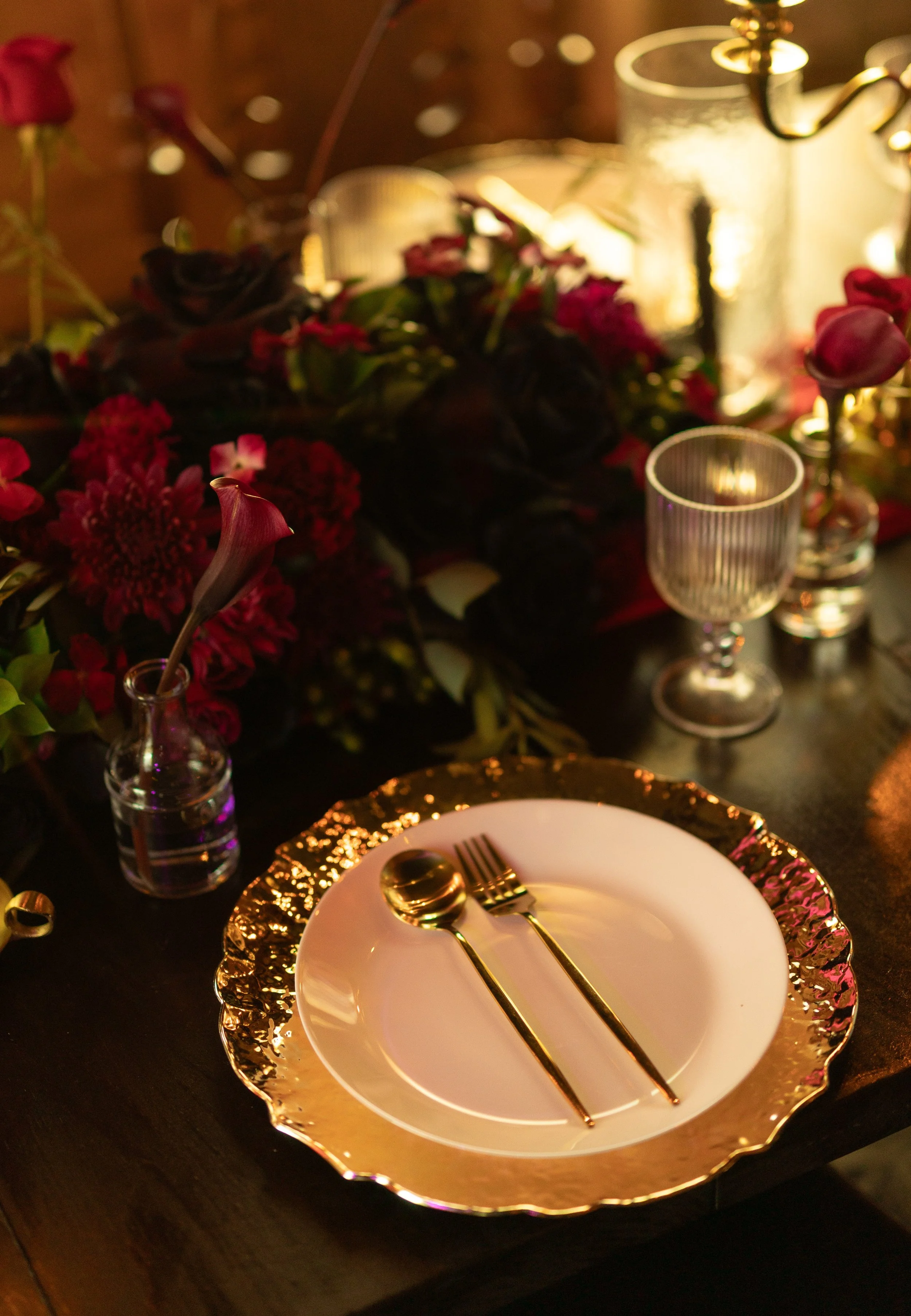 Table setting with a white plate, gold cutlery, a gold-rimmed charger, and a gold-rimmed glass. In the background, there is a flower arrangement with red and pink flowers, and a candle holder with lit candles, creating a warm, intimate ambiance.