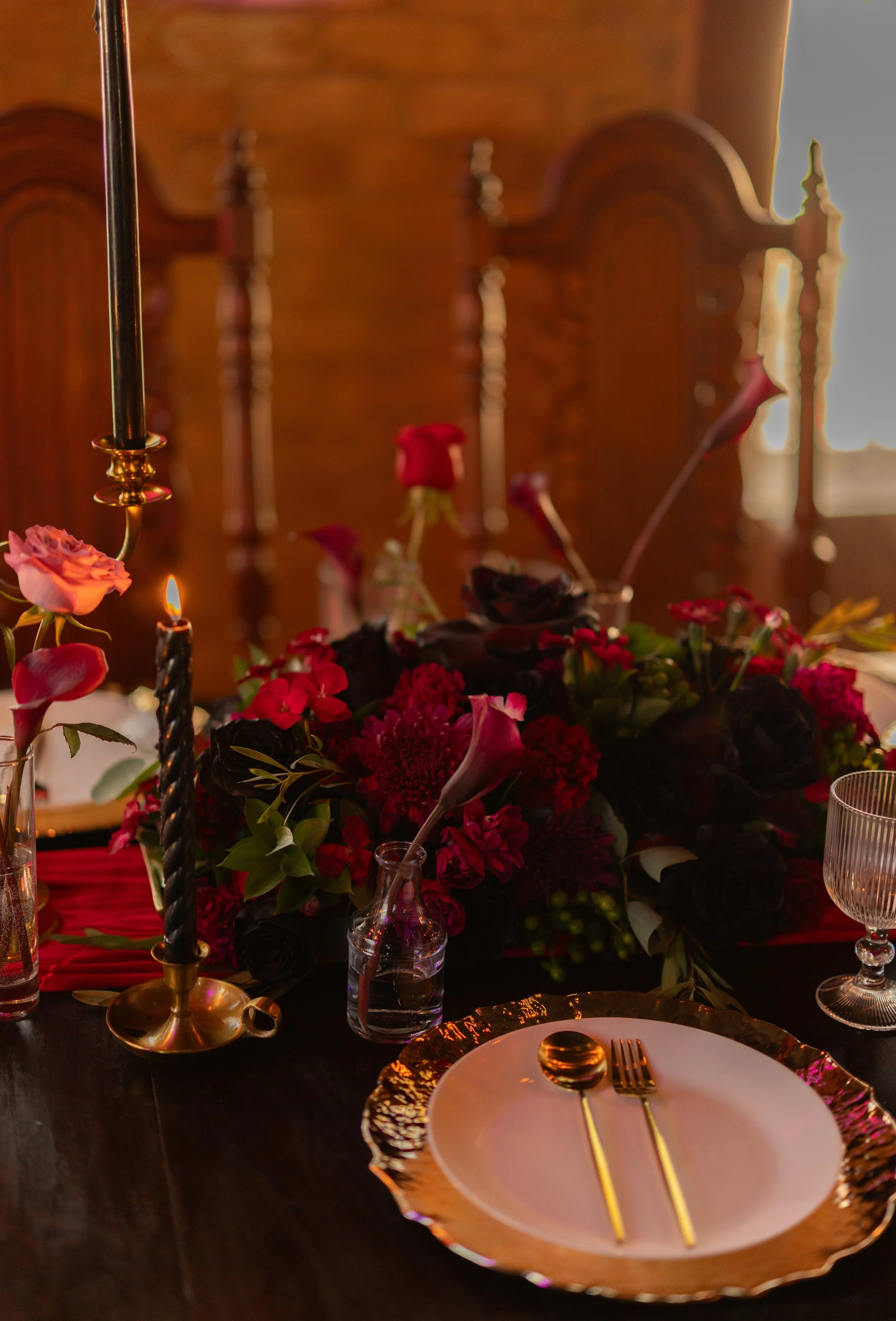 A table setting with a white plate, gold-edged charger, gold fork and spoon, surrounded by a floral centerpiece with red and dark-colored flowers, tall black candles in gold holders, and a background of wooden furniture.