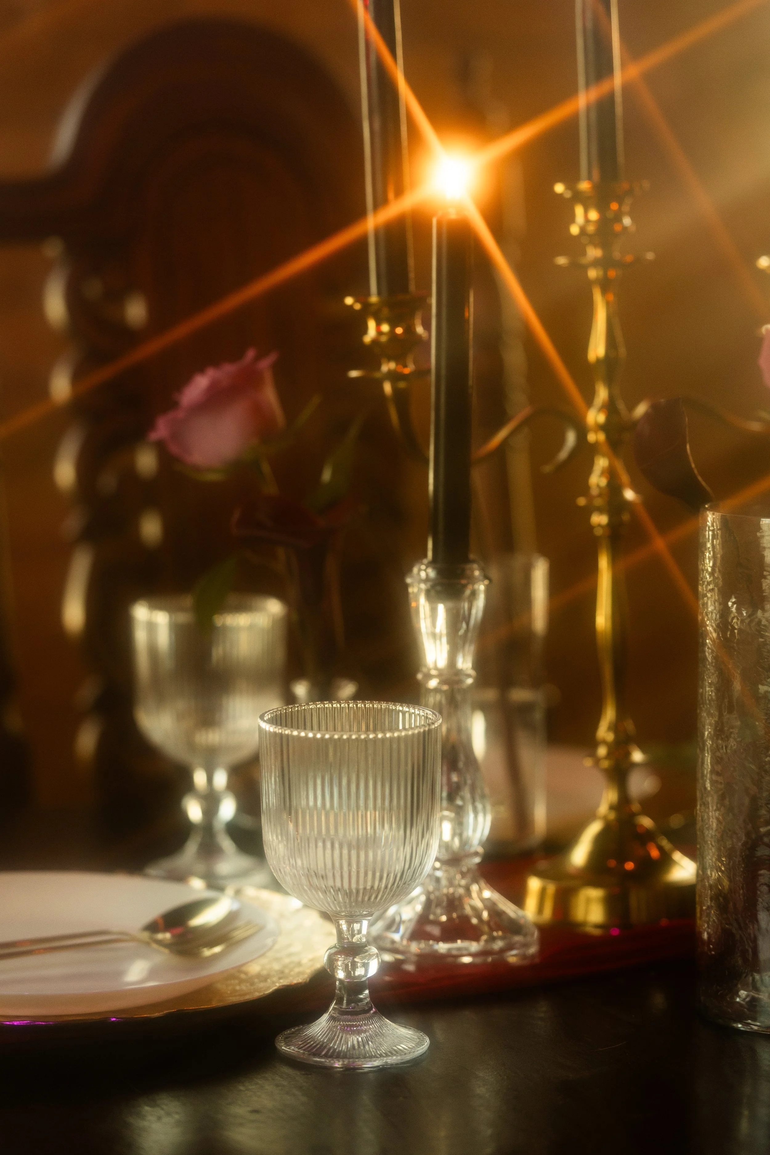 A close-up of a dining table set with glassware, including wine glasses, candle holders, and a pink flower in a glass vase, with warm lighting and a bright light source creating lens flare.