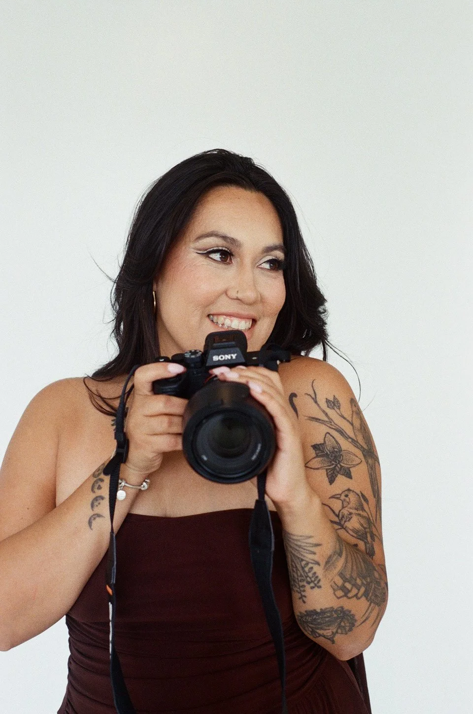 A woman with black hair and tattoos holding a Sony camera, smiling, against a plain white background.