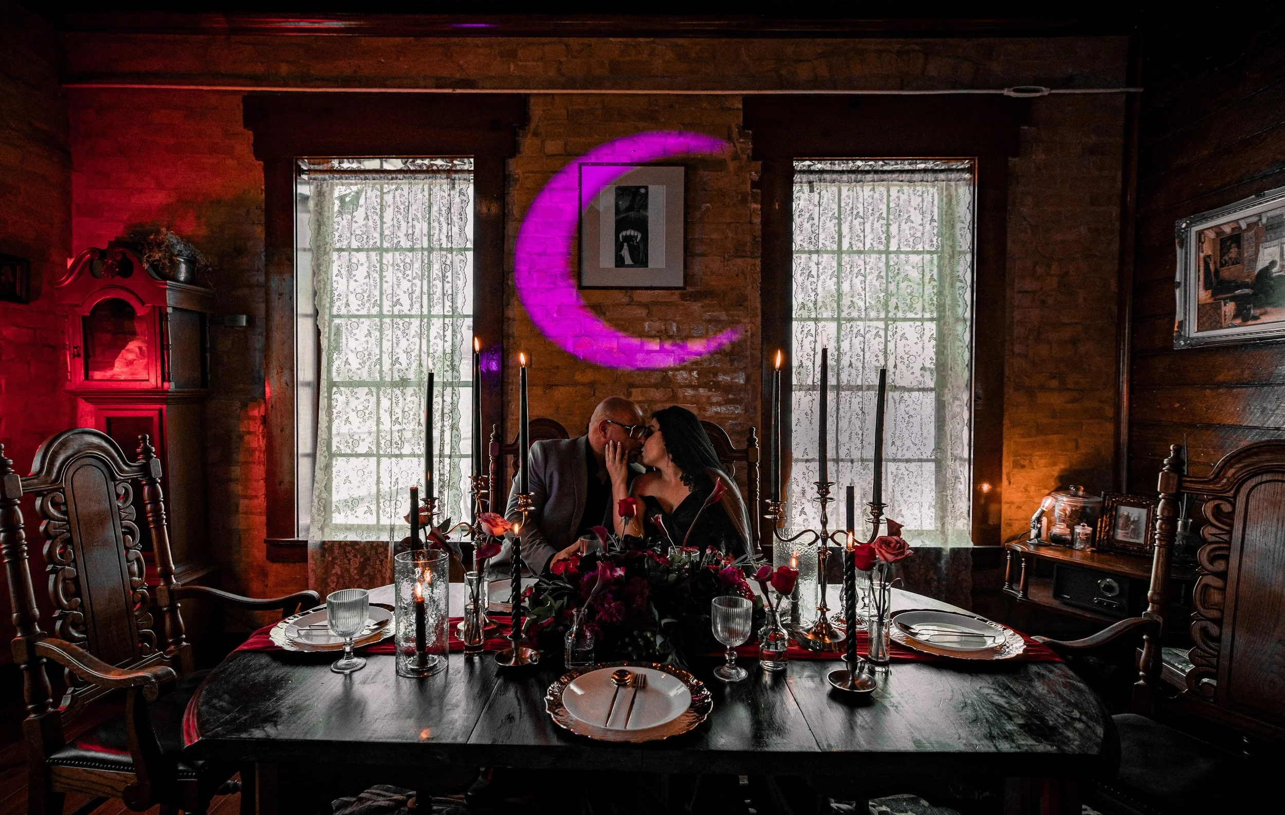 A romantic couple is seated closely together at a dinner table decorated with flowers and candles, inside a cozy wooden room with lace curtains and artwork on the walls, illuminated by purple moon-shaped neon light.