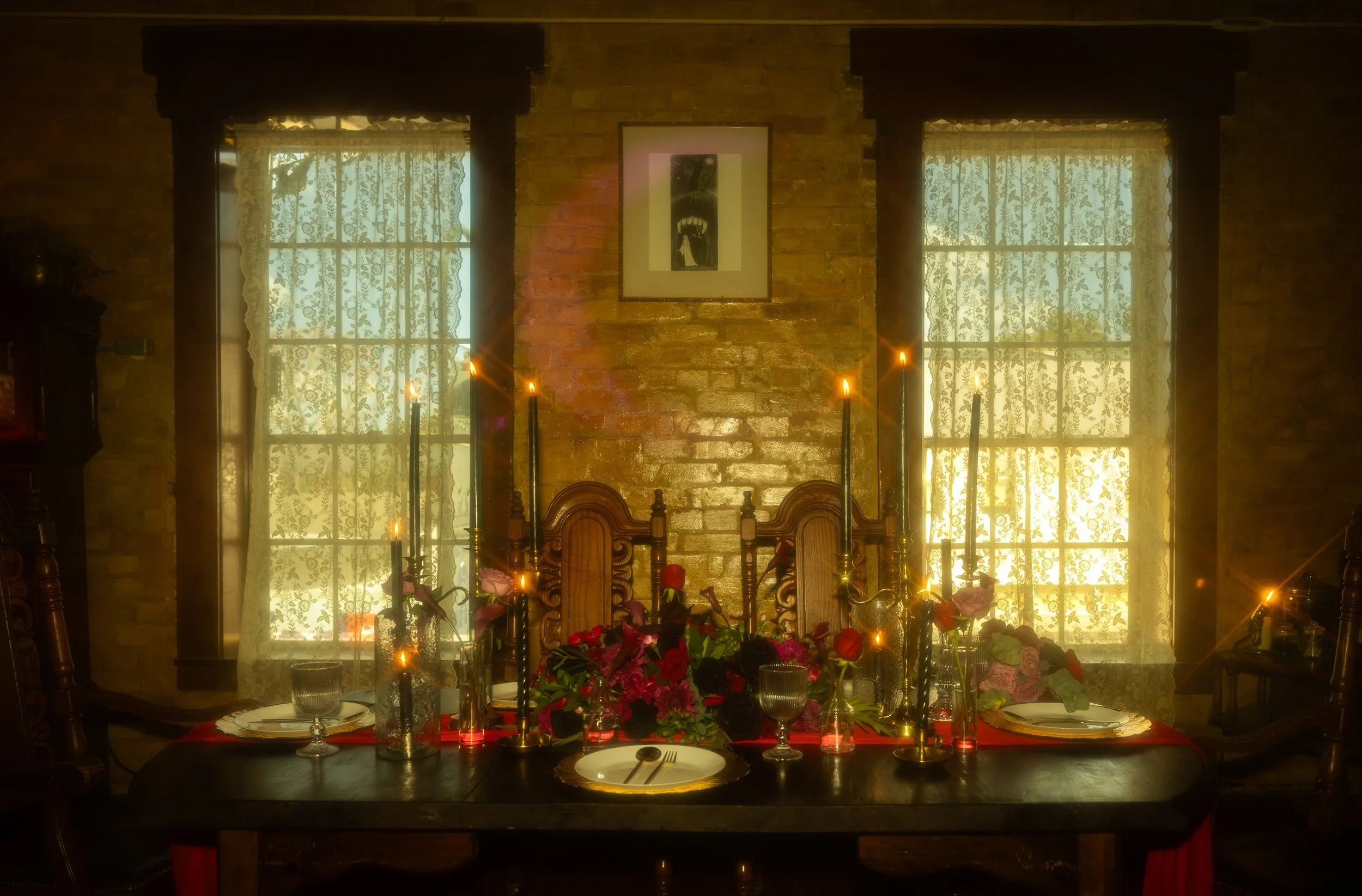 A dining room with a table set for a meal, decorated with flowers and candles, in front of two windows with lace curtains, and a brick wall with a framed picture.