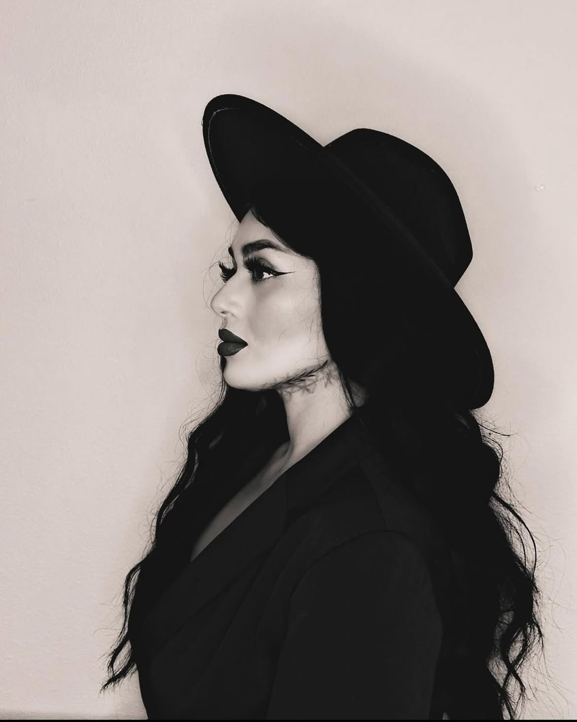 Black and white photo of a woman in profile wearing a wide-brimmed hat, with dramatic makeup and long wavy hair.