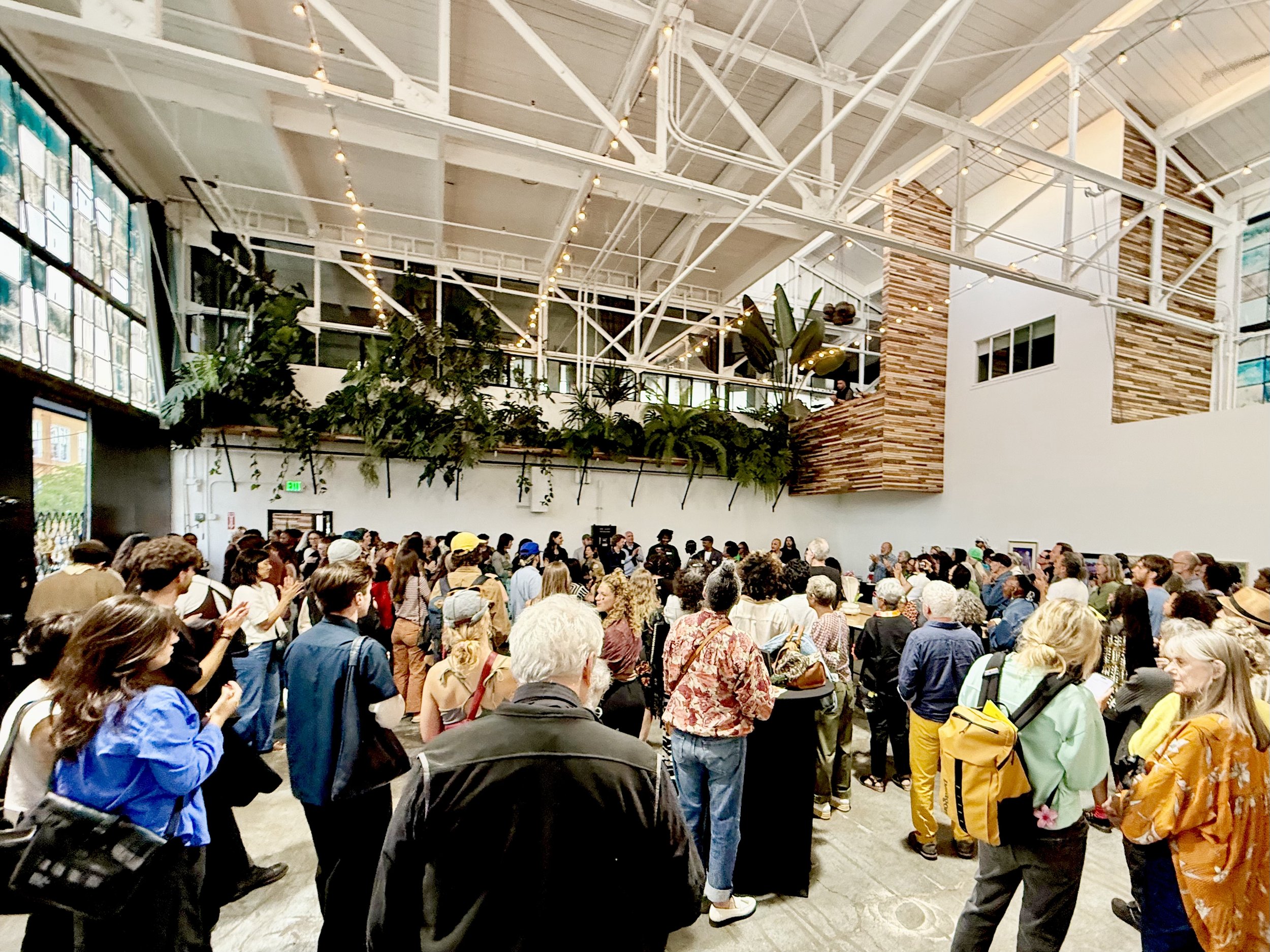 Crowd of visitors at an open studio event in a bright warehouse art space with plants and string lights