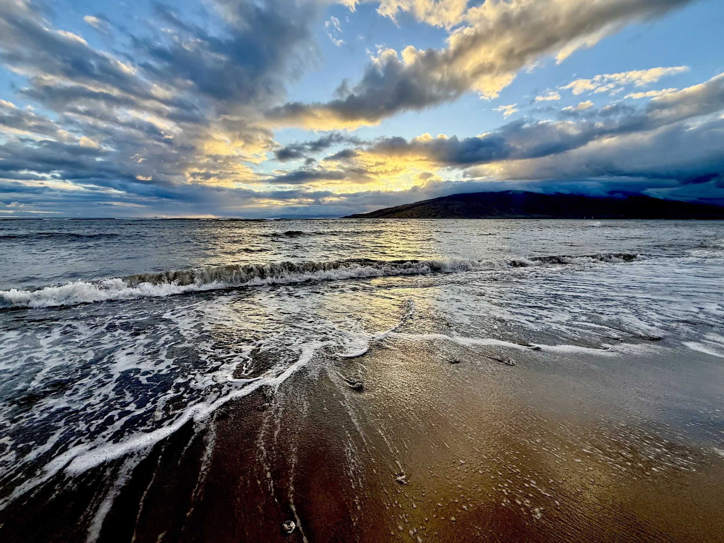 Waves washing onto a sandy shoreline at sunset in Maui, Hawaii, with a mountain on the horizon — the ocean that inspires Susan Brady's art