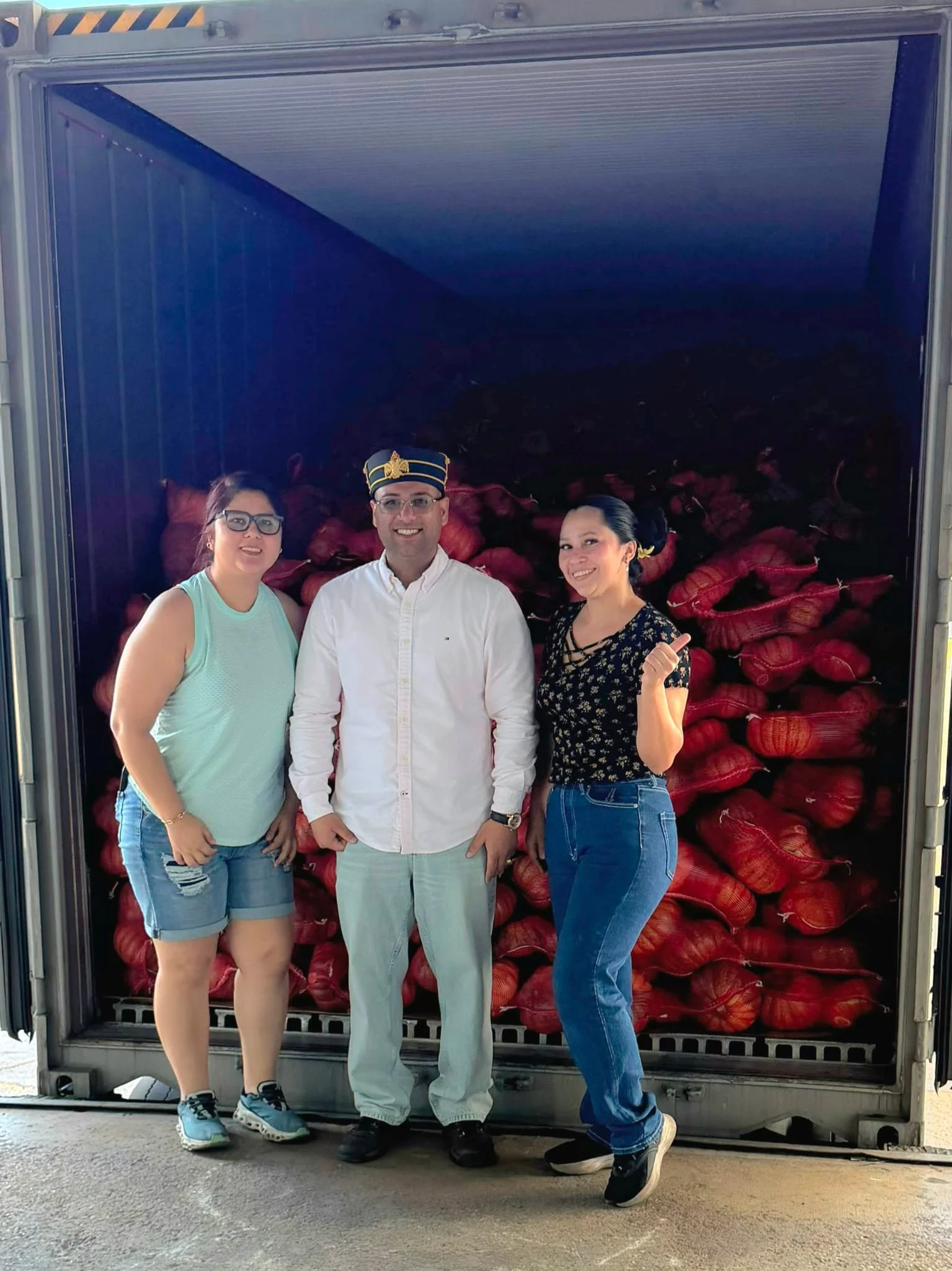 Three people standing in front of a large container filled with red onions. The person in the middle is wearing a white shirt and a hat with gold accents. The female on the left is wearing glasses, a sleeveless turquoise top, and denim shorts. The female on the right is wearing a dark floral top and jeans, giving a thumbs-up.